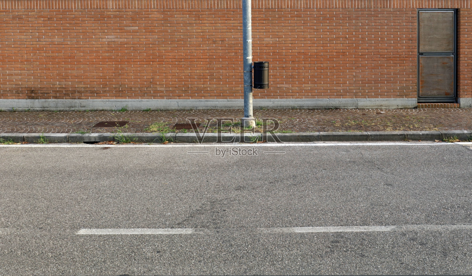 人行道上灯杆上的黑色垃圾桶。房子后面有砖墙。城市道路在前方。照片摄影图片