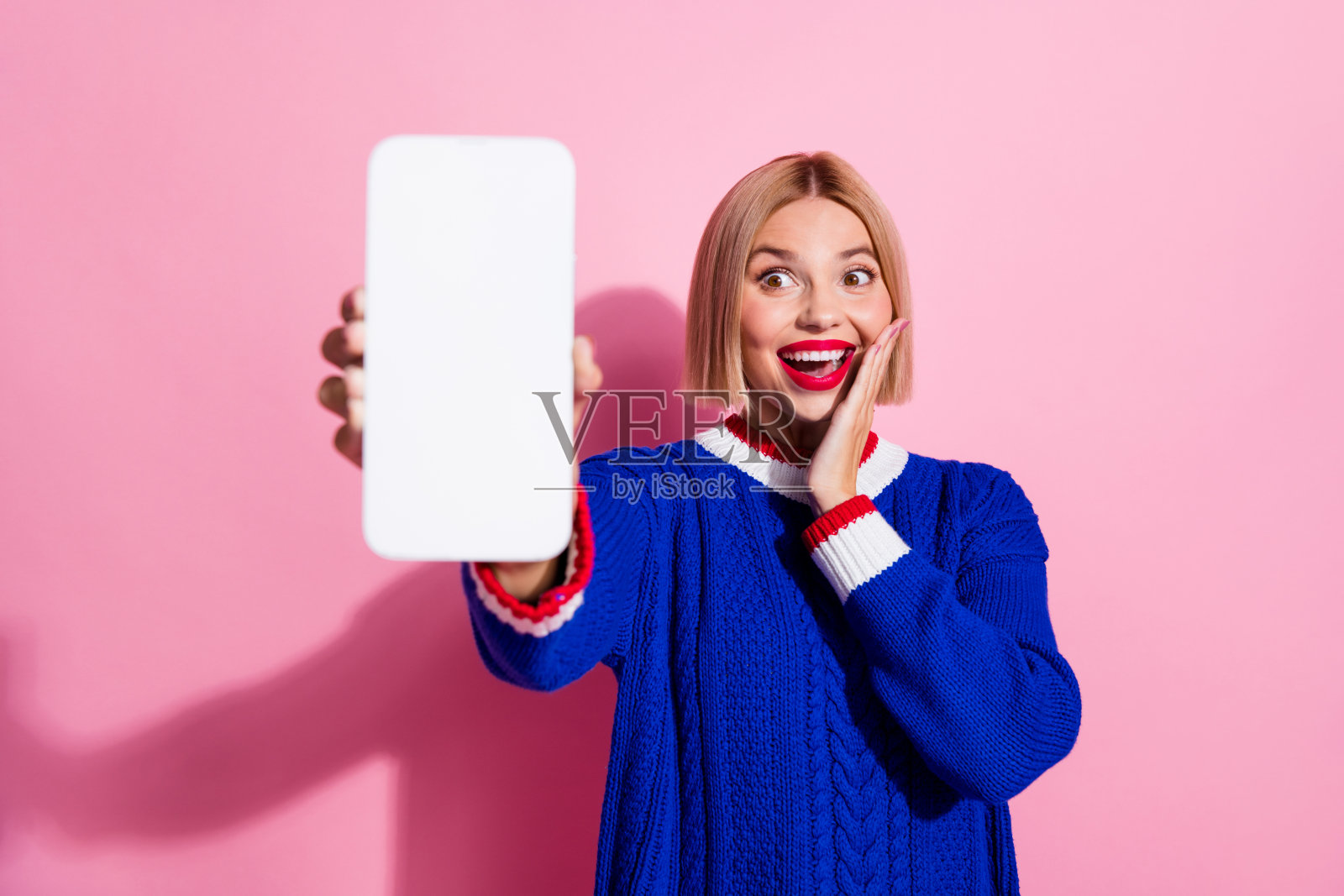 年轻震惊的金发波波头女士的肖像，穿着蓝色套头衫，手持显示器，触摸脸颊，看到它孤立在粉红色的背景上照片摄影图片