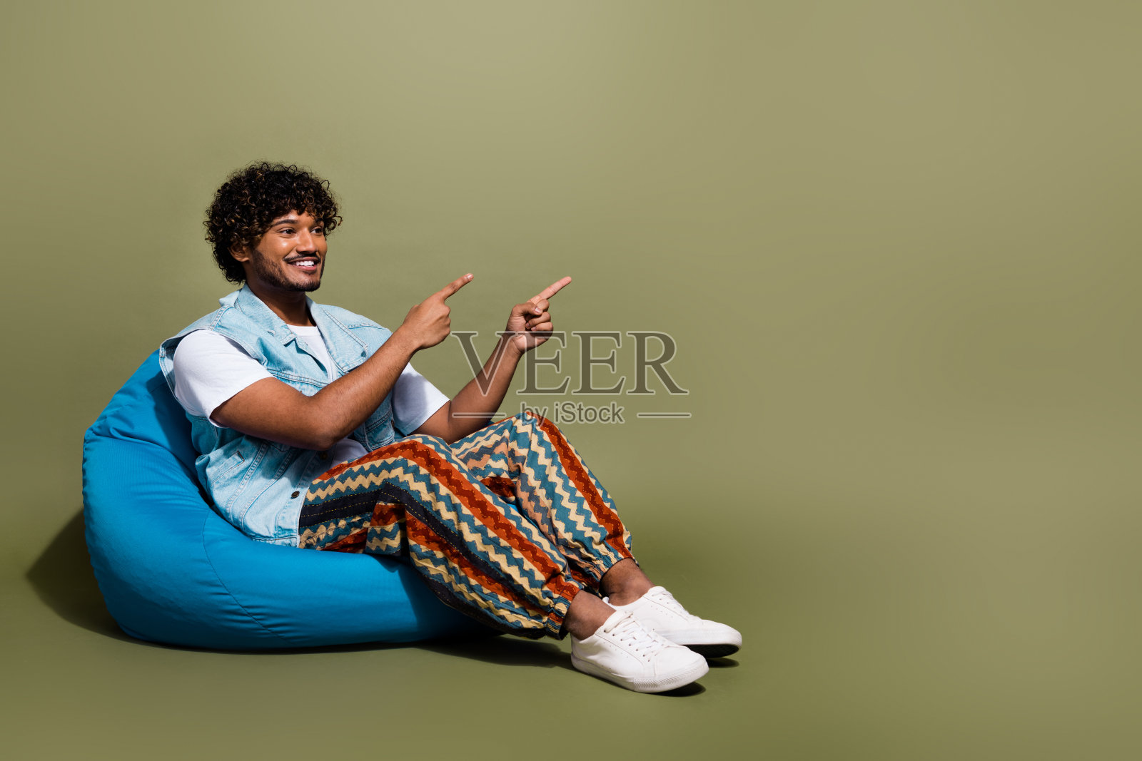 全尺寸照片英俊的年轻人坐在豆袋点看空空间穿时髦的牛仔裤装孤立的卡其色背景照片摄影图片