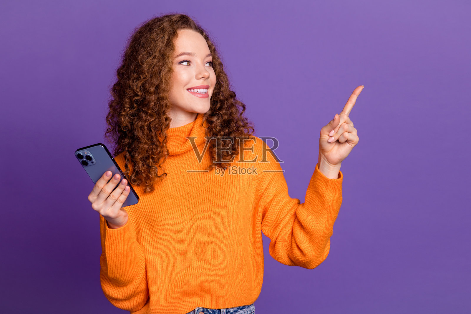 照片中可爱的年轻女孩拿着手机直接手指空着，穿着橙色的套头衫孤立在紫罗兰色的背景上照片摄影图片