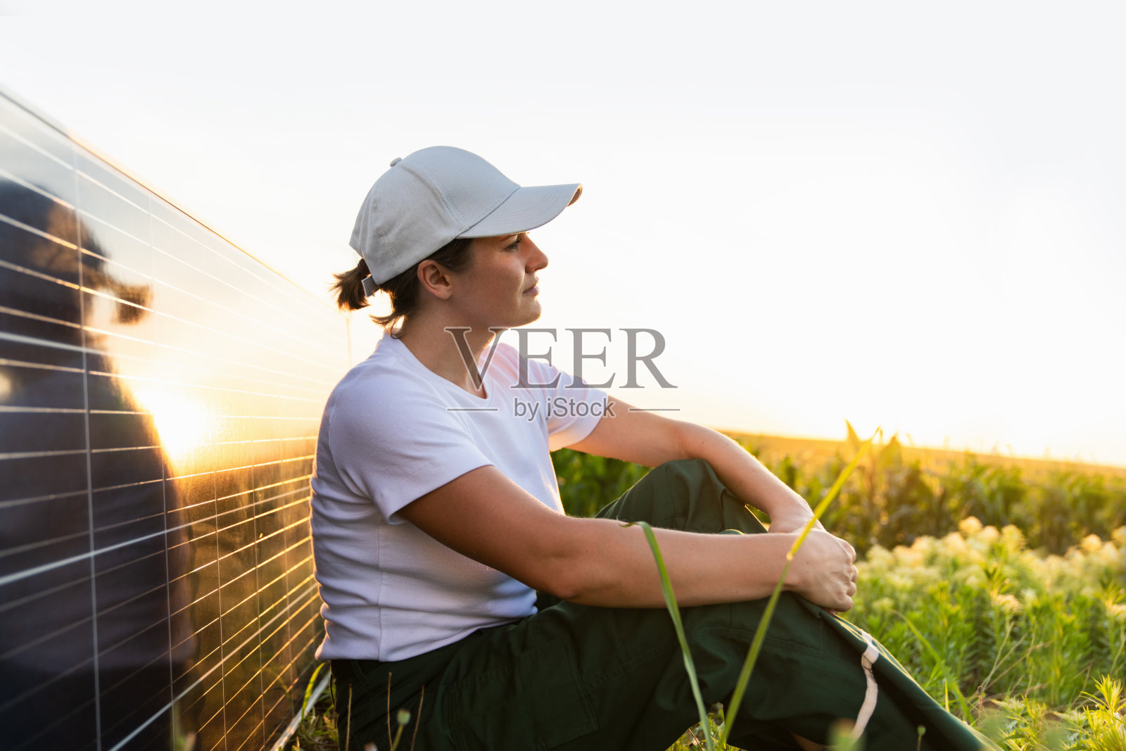 日落时分，戴着白色帽子的妇女坐在太阳能电池板旁。背景是风力涡轮机照片摄影图片