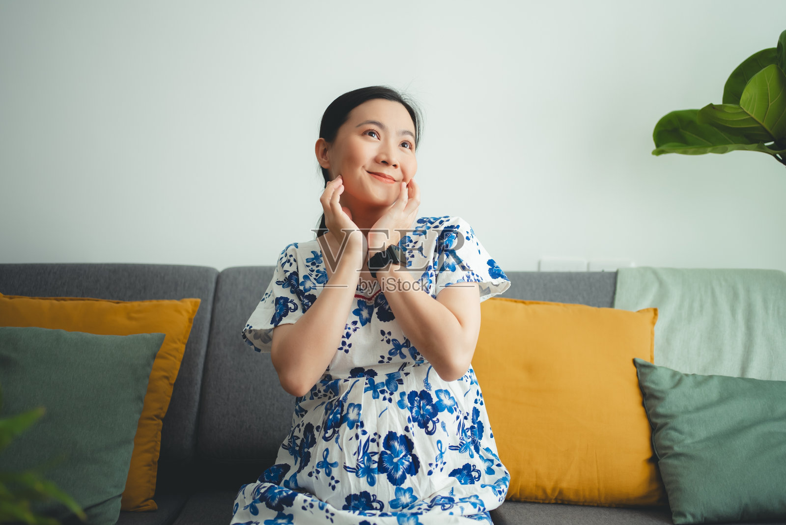 亚洲孕妇坐在家里客厅的沙发上，开心地笑着，有想法。照片摄影图片