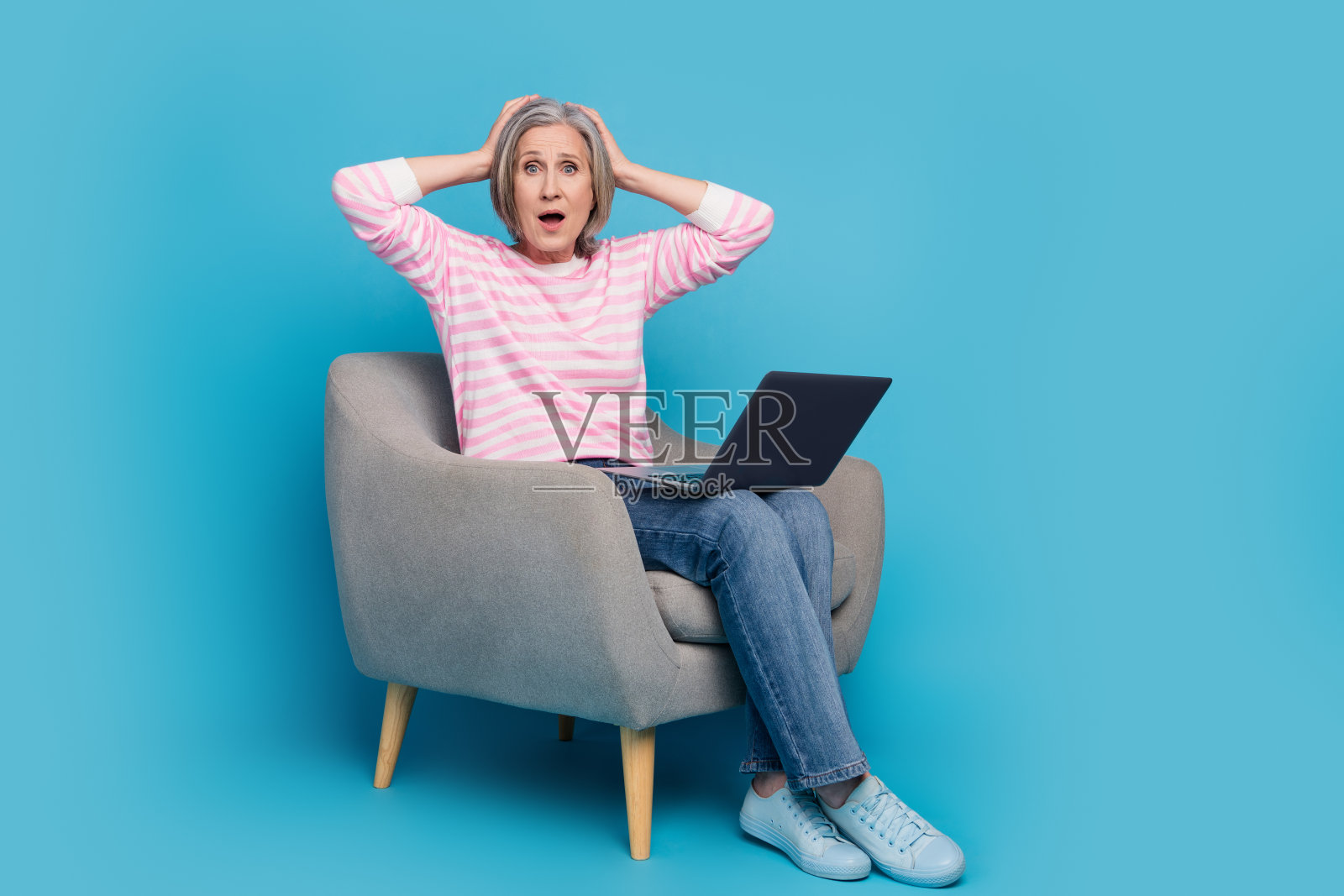 漂亮的老年妇女的全身照片，坐在扶手椅上网本，害怕穿着时髦的条纹服装，孤立在蓝色的背景照片摄影图片