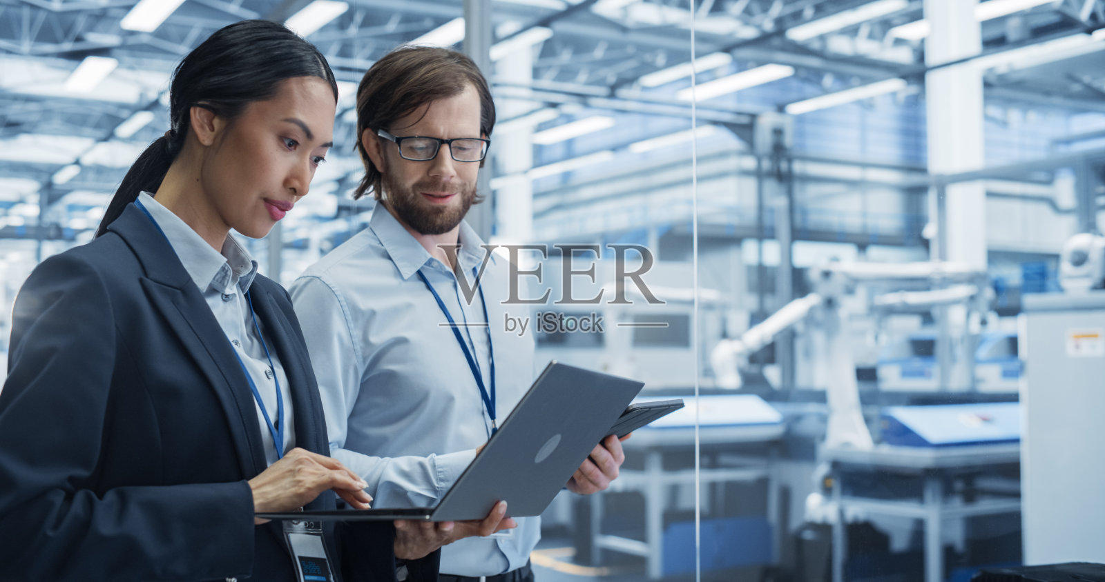 经验丰富的男女工程师站在机构房间里，使用电脑设备讨论现代自动化电子制造工厂的生产，工厂配备了机器人手臂照片摄影图片