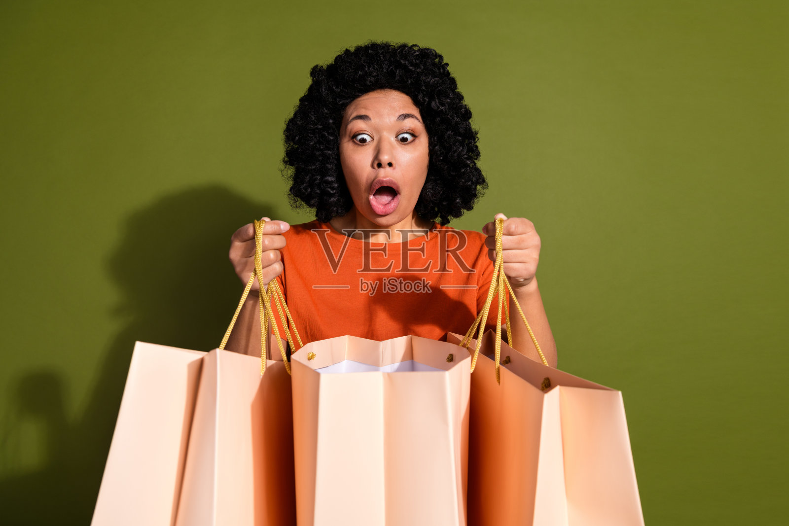 照片中漂亮的年轻女子看着商店的袋子，穿着橙色的t恤，孤立在卡其色的背景上照片摄影图片