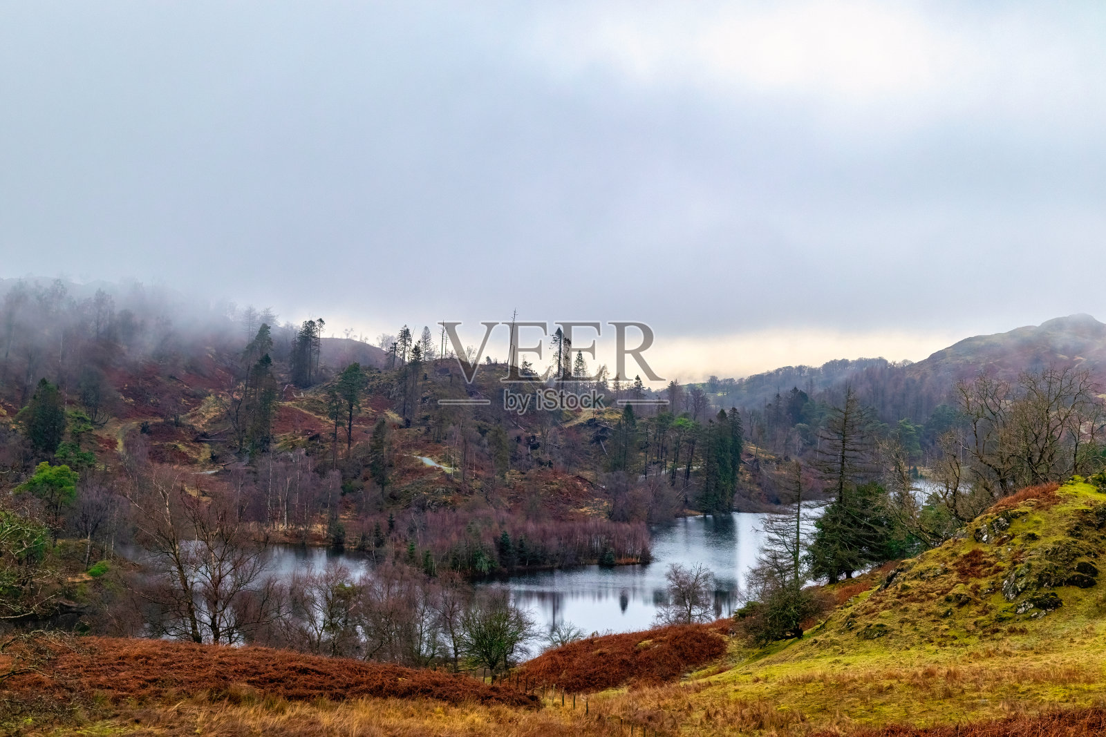 雾气蒙蒙的风景，宁静的湖泊被秋天的森林环绕，连绵起伏的丘陵在多云的天空下，英格兰湖区。照片摄影图片