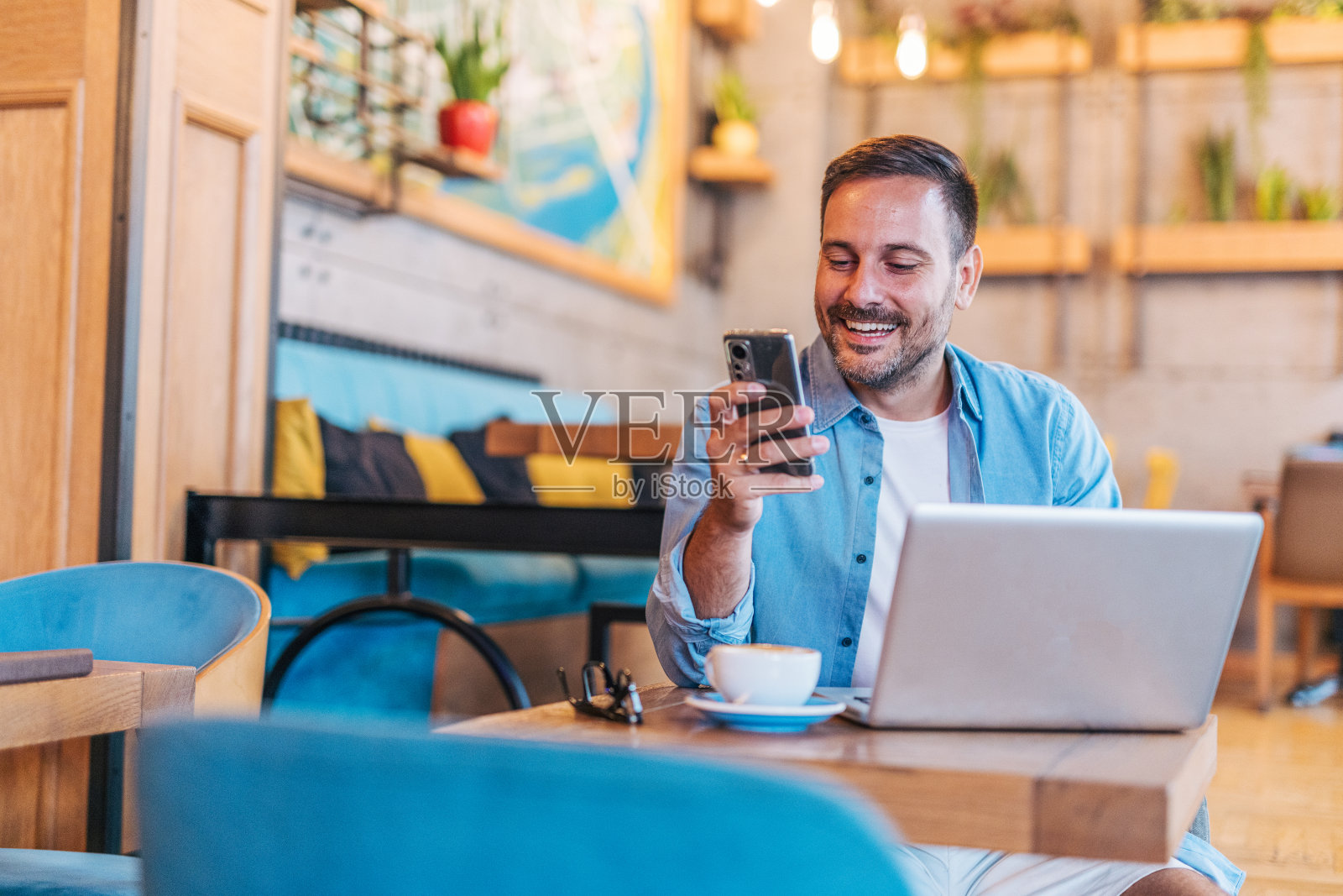 快乐英俊的中年男子在咖啡馆享受宁静的咖啡休息时间，在使用笔记本电脑的同时使用智能手机照片摄影图片