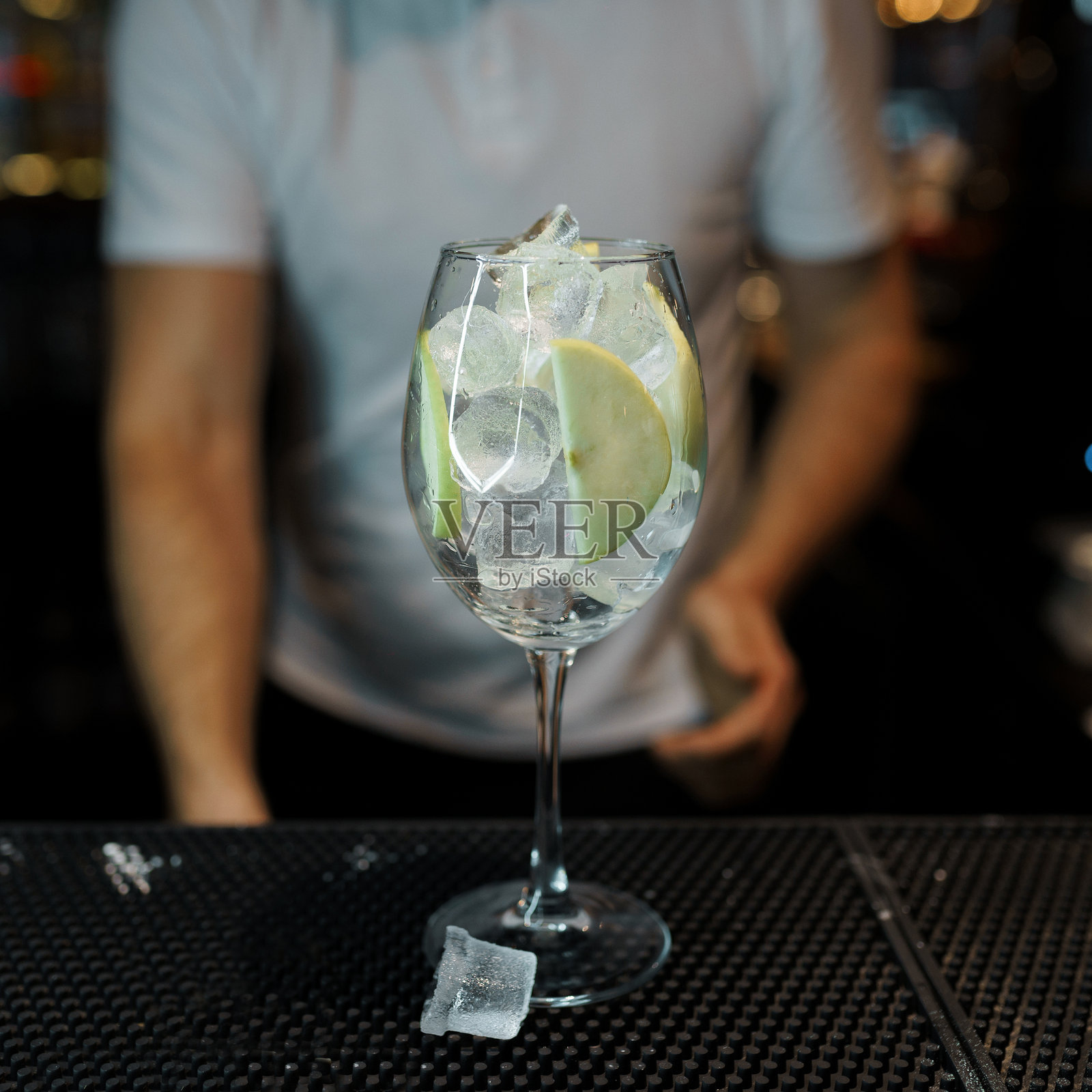 吧台上放着冰和新鲜苹果的鸡尾酒，背景是穿着白色t恤的酒保。为好心情做一杯美味的鸡尾酒照片摄影图片