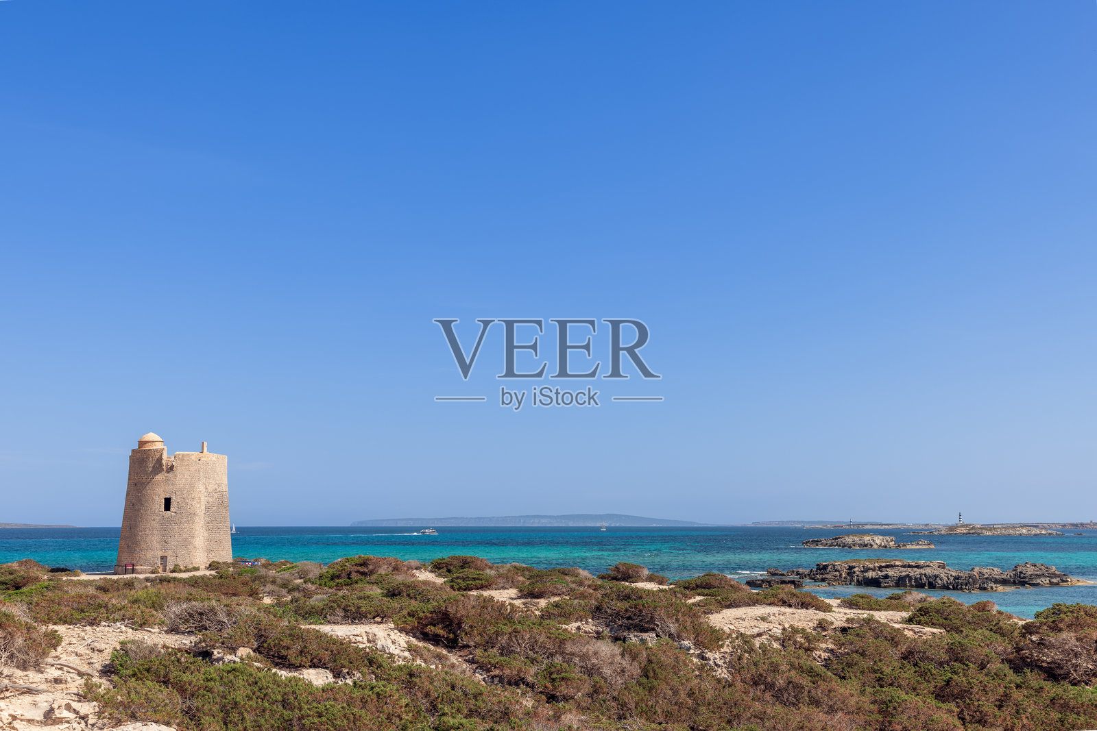 位于伊比沙岛海岸的Torre de ses Portes矗立在绿松石般的大海和清澈的蓝天下，周围是稀疏的海岸植被，将历史与宁静的自然美景融为一体照片摄影图片