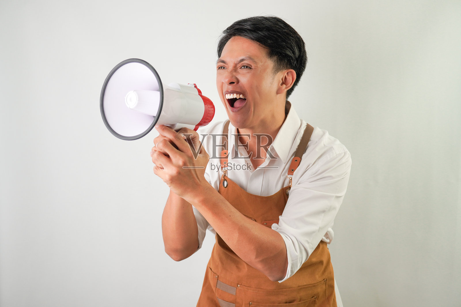 穿着卷起的白袖衬衫，系着棕色围裙的兴奋的年轻亚洲人带着微笑的表情拿着扩音器。对着扩音器尖叫。隔离在白色背景上照片摄影图片