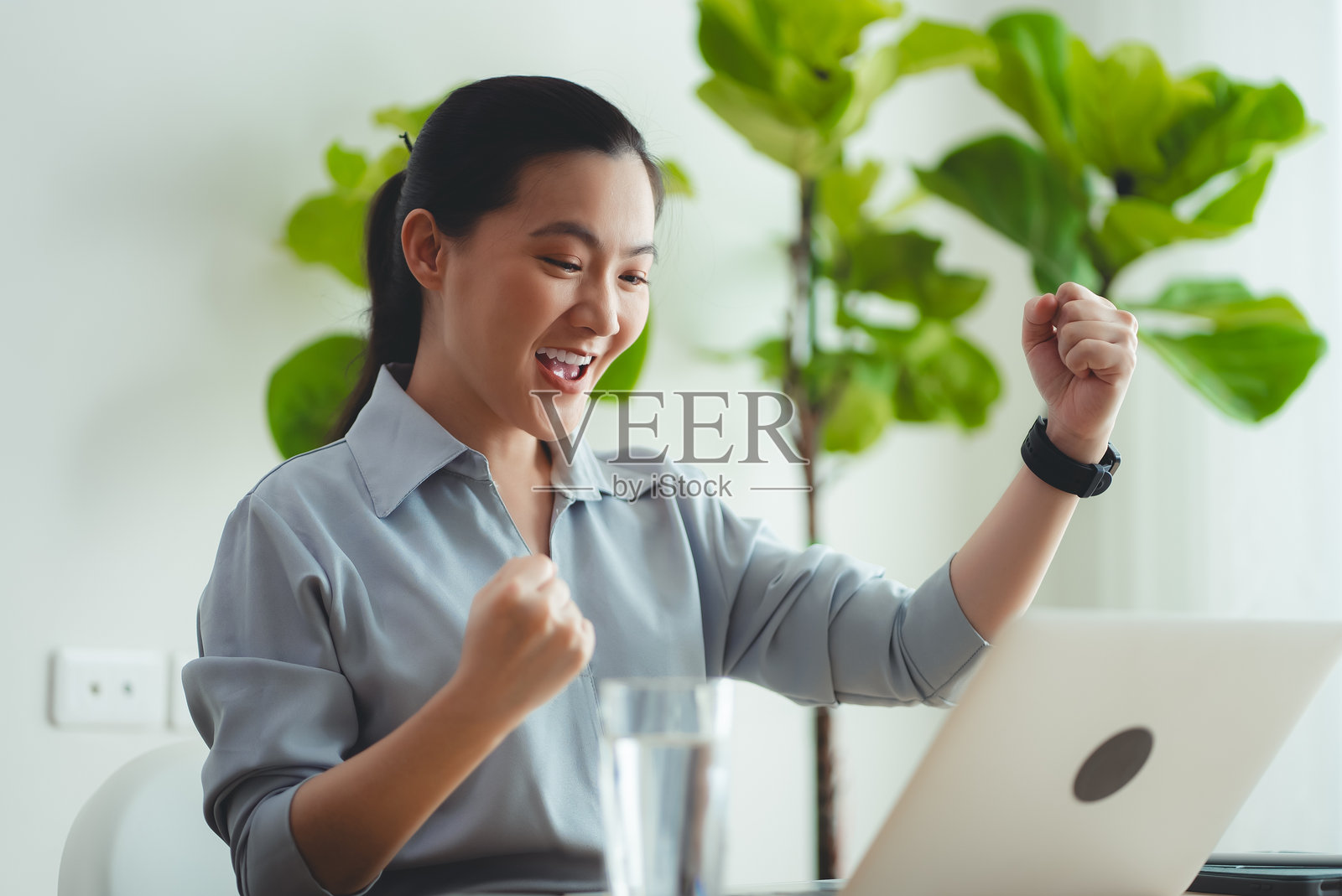 亚洲女性坐在家里办公室里，用着笔记本电脑，开心兴奋，做出了一个获胜的手势。照片摄影图片