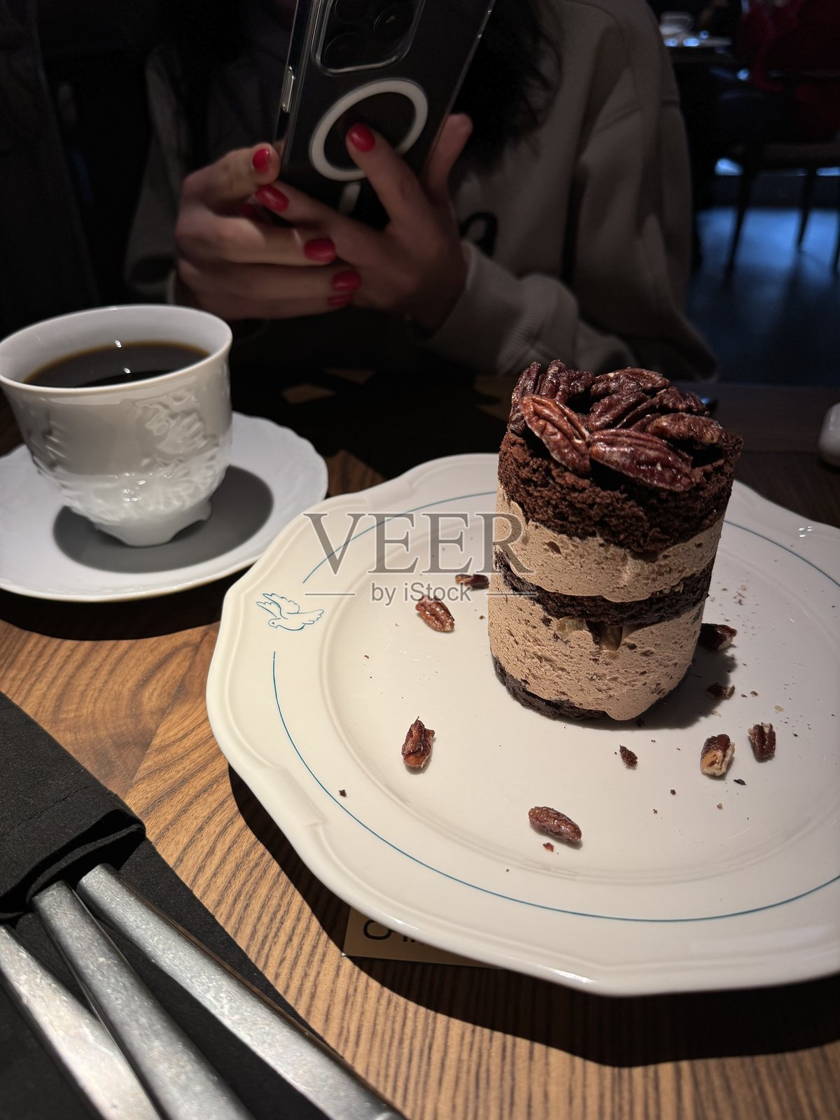 白色盘子里分层的巧克力甜点和山核桃的特写，配上一杯黑咖啡。一个涂着红指甲的女人拿着智能手机，在一个舒适的咖啡馆里拍下了这一幕。照片摄影图片