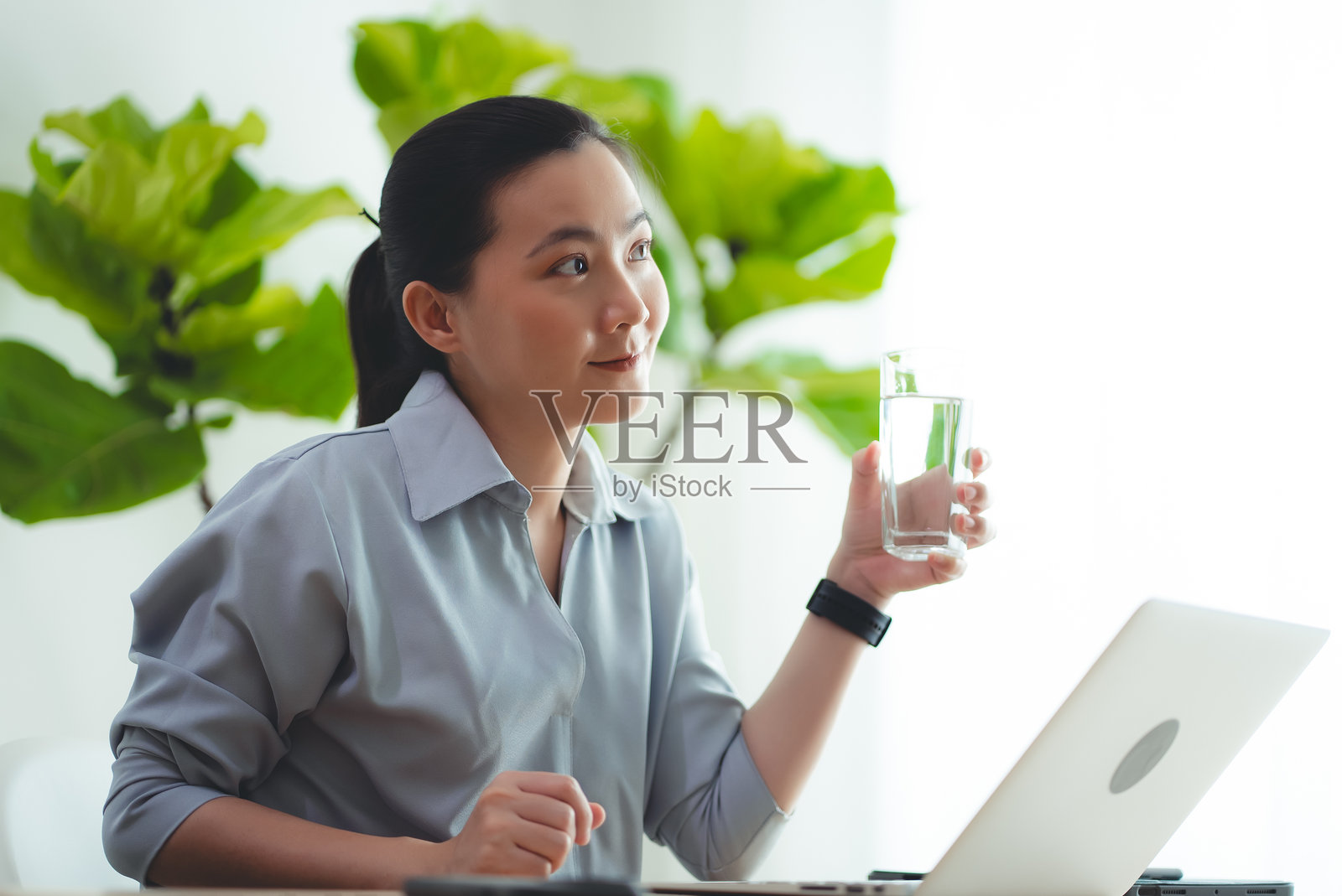 亚洲女性坐在家里办公室边喝水边用笔记本电脑休息。照片摄影图片
