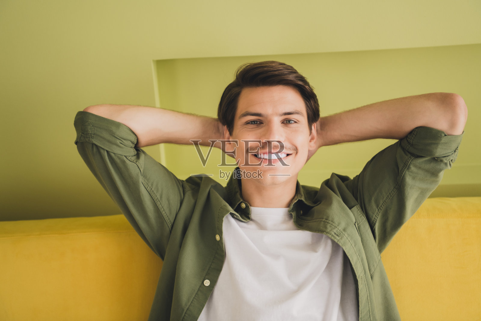 梦幻般的有吸引力的好男人坐在舒适的沙发上，在头后面抱着手臂，在室内享受现代的室内住宅照片摄影图片