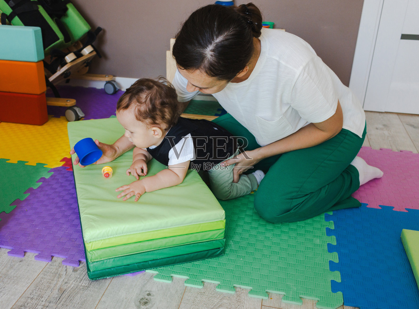 一个肌肉无力的男孩穿着脊柱紧身衣，在治疗师的帮助下进行康复训练，使用彩色垫子和辅助装备照片摄影图片