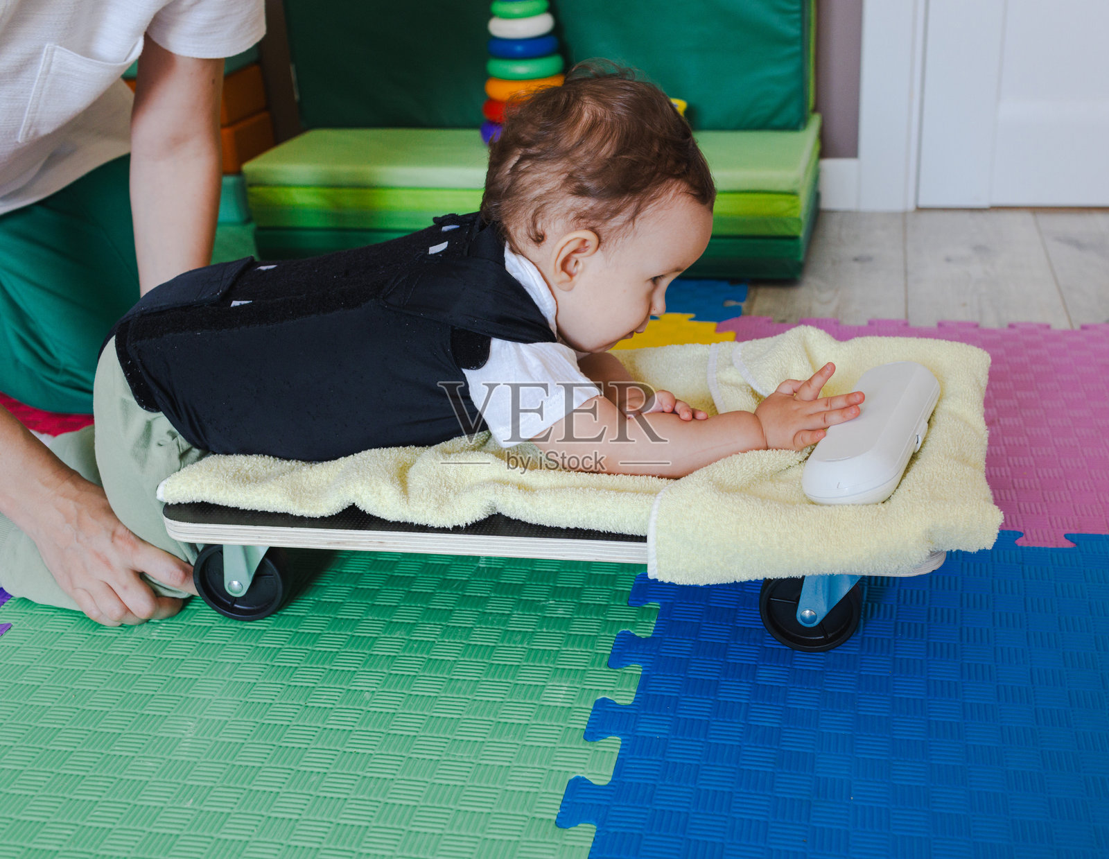 在康复诊所，中枢神经系统受损的男童穿着脊柱紧身衣与物理治疗师一起使用轮式平台学习爬行照片摄影图片