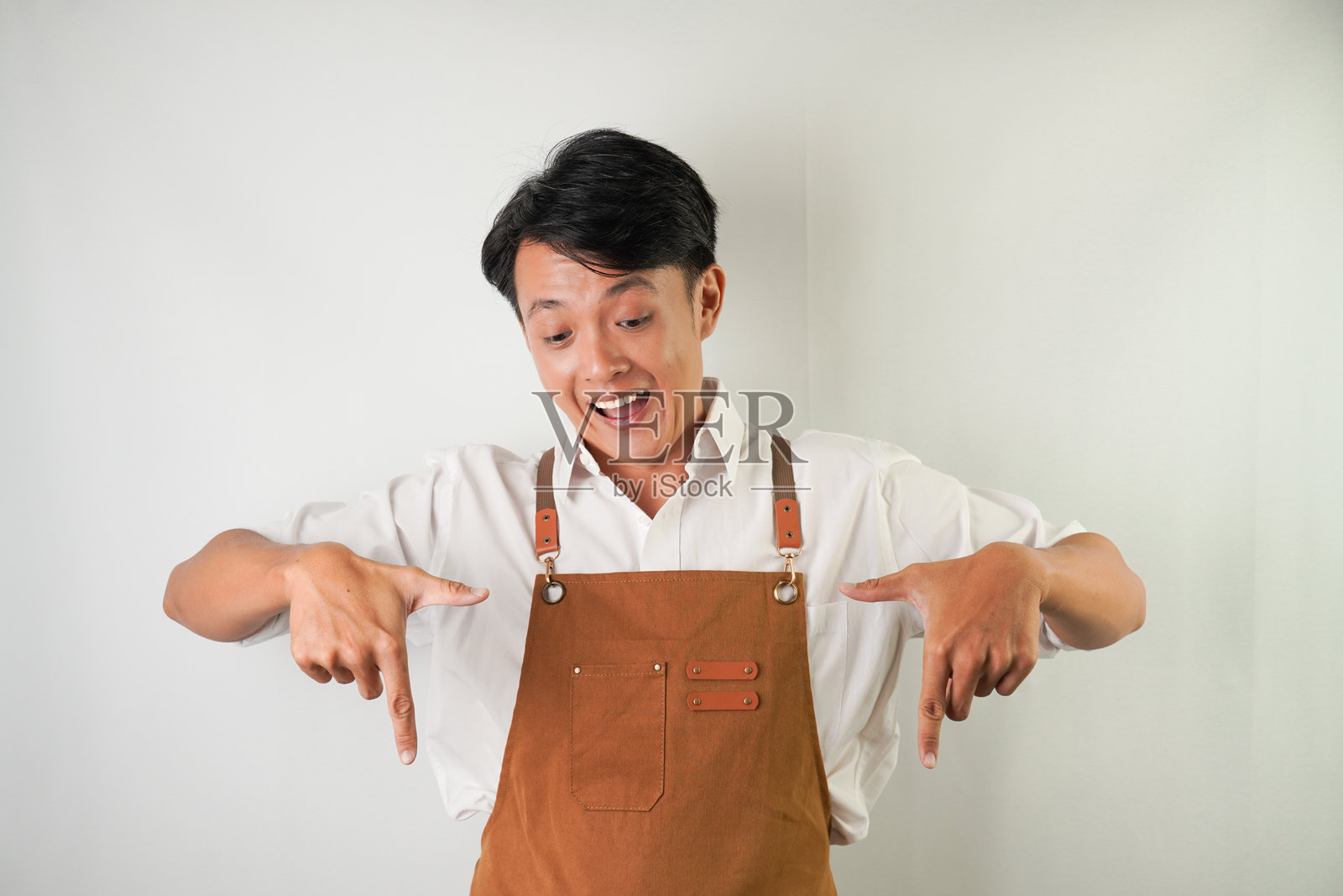 穿着卷起的白袖衬衫，系着棕色围裙的兴奋的年轻亚洲人正带着微笑的表情向下指着。隔离在白色背景上照片摄影图片