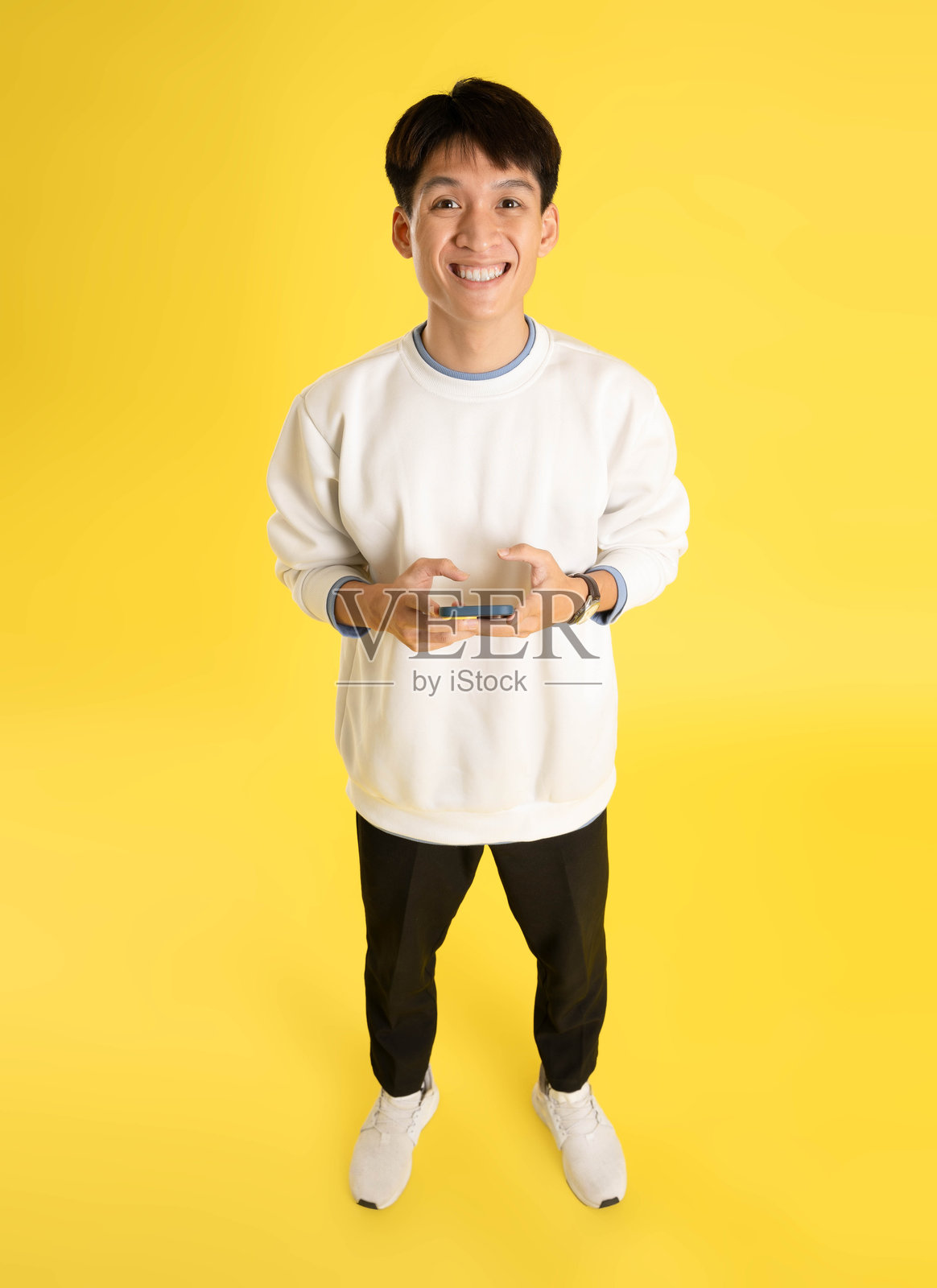 黄色背景上，一名年轻的亚洲男子全身穿着毛衣，正在使用手机照片摄影图片