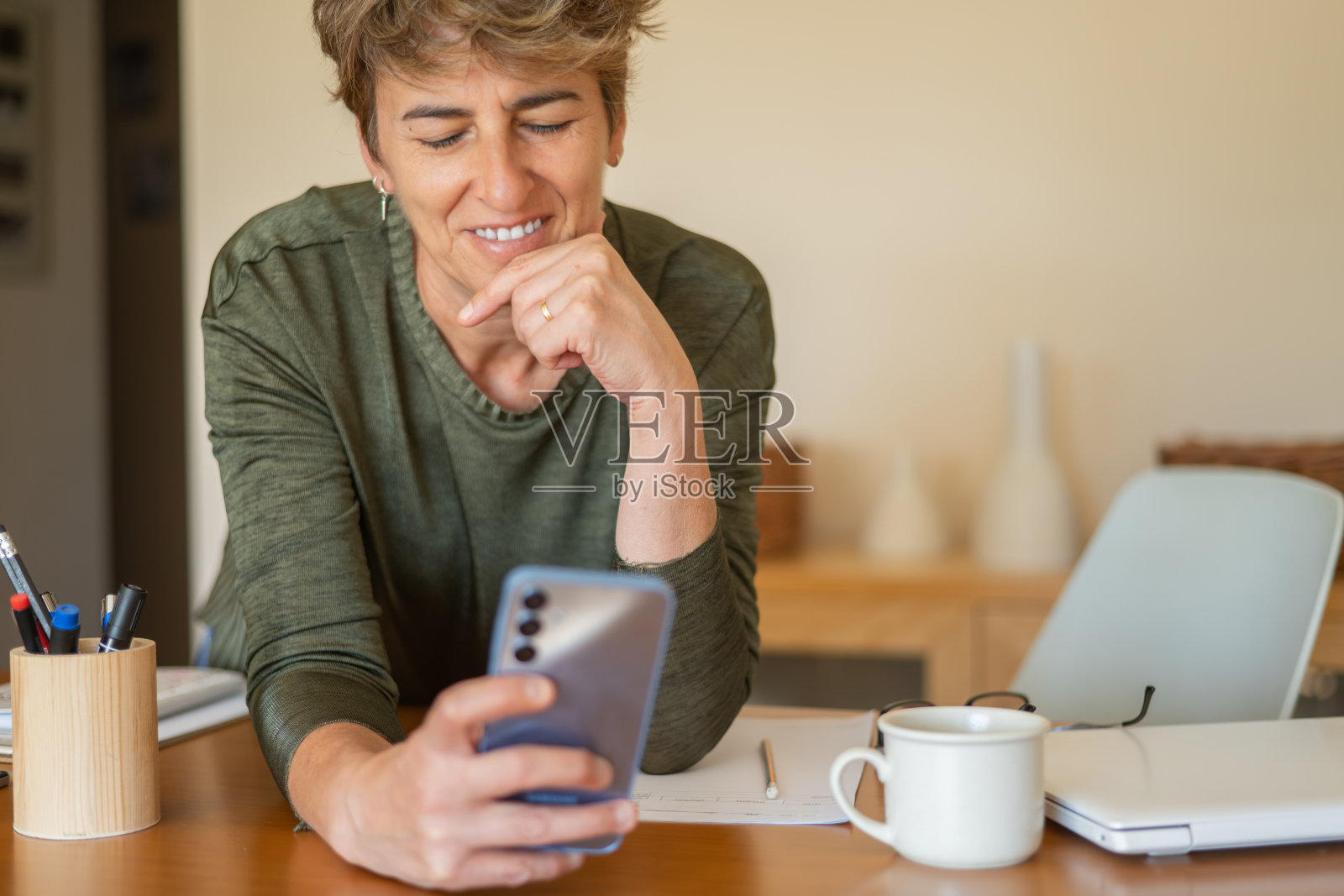 微笑的成熟的女性在工作场所或家中使用智能手机，好消息，享受交换信息，从工作中分心，花时间上网，与朋友或家人聊天，看新闻。照片摄影图片