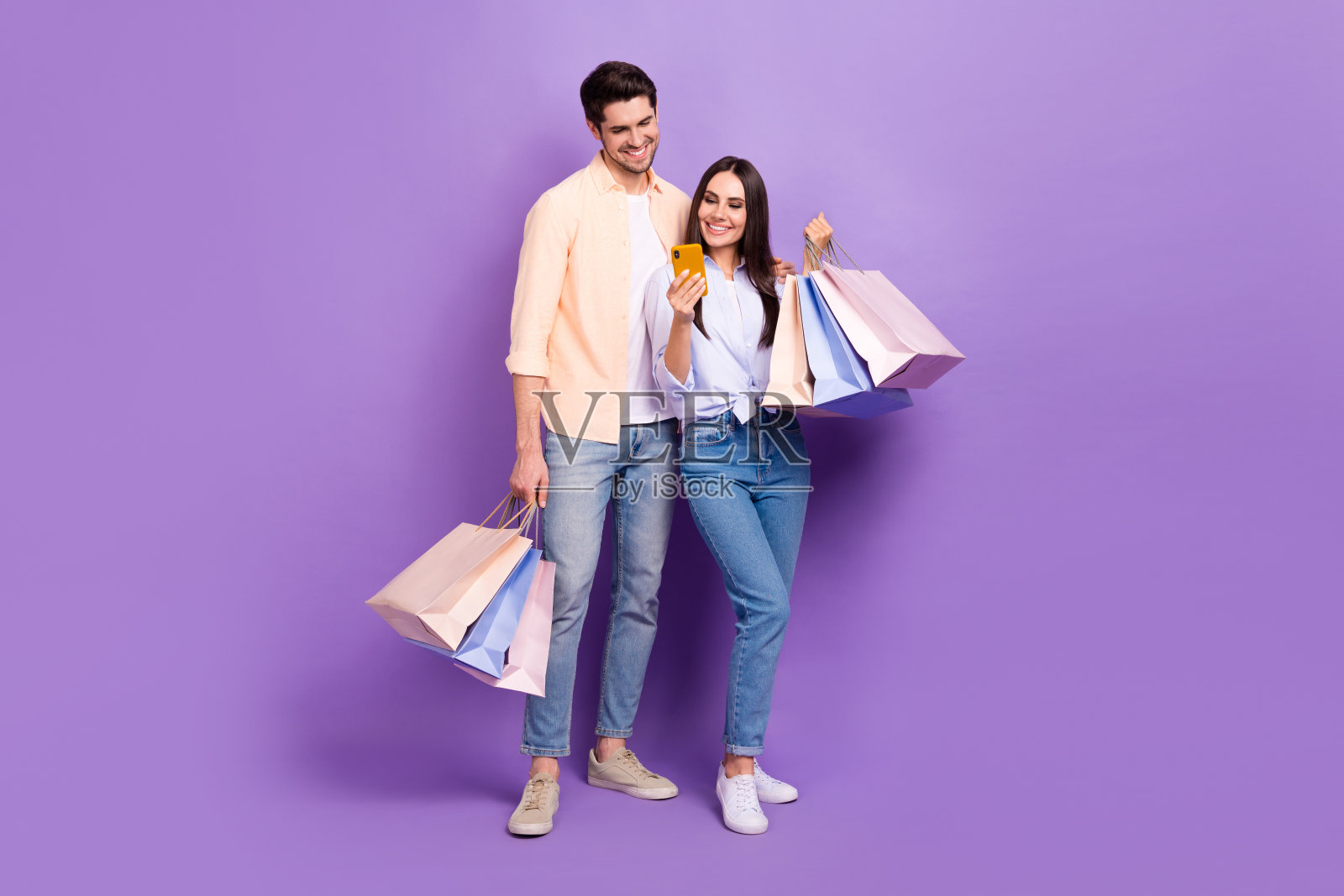 全尺寸照片，两个有吸引力的积极的人拥抱拿着购物袋使用智能手机孤立的紫色背景照片摄影图片