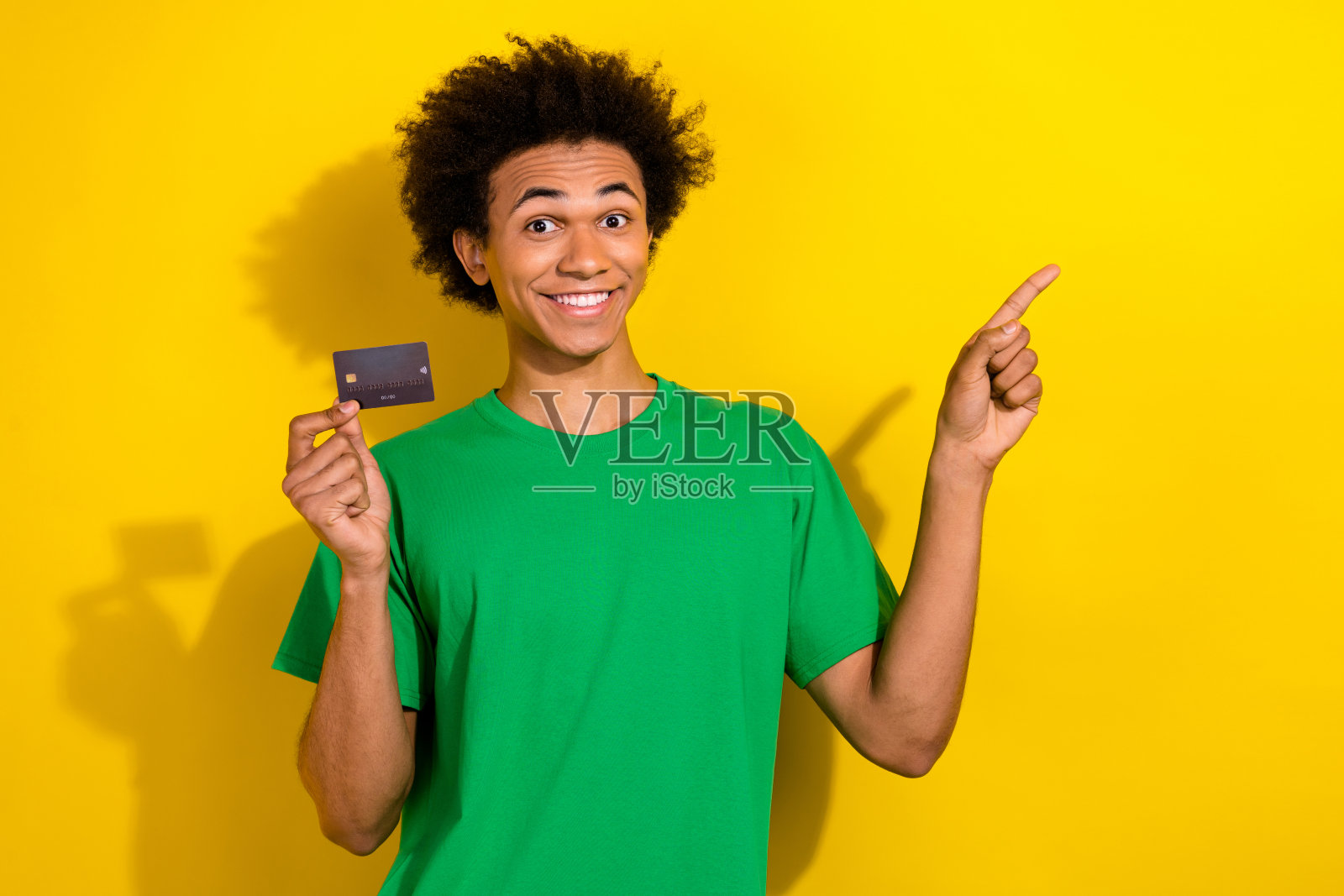 照片中开朗积极的小伙子穿着绿色t恤，手持银行卡指着空旷的空间孤立的黄色背景照片摄影图片