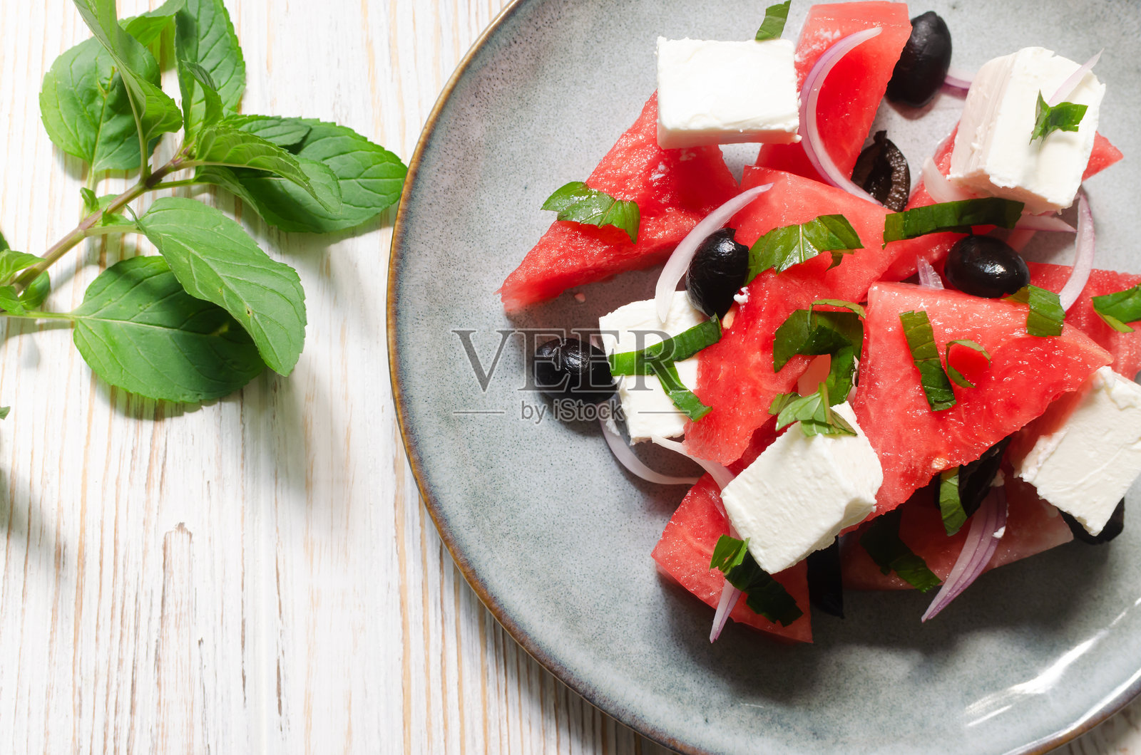 白色木桌上，灰色盘子里放着夏日沙拉，里面有西瓜、羊乳酪、橄榄和薄荷。健康饮食的概念。素食,素食者。水平方向。照片摄影图片