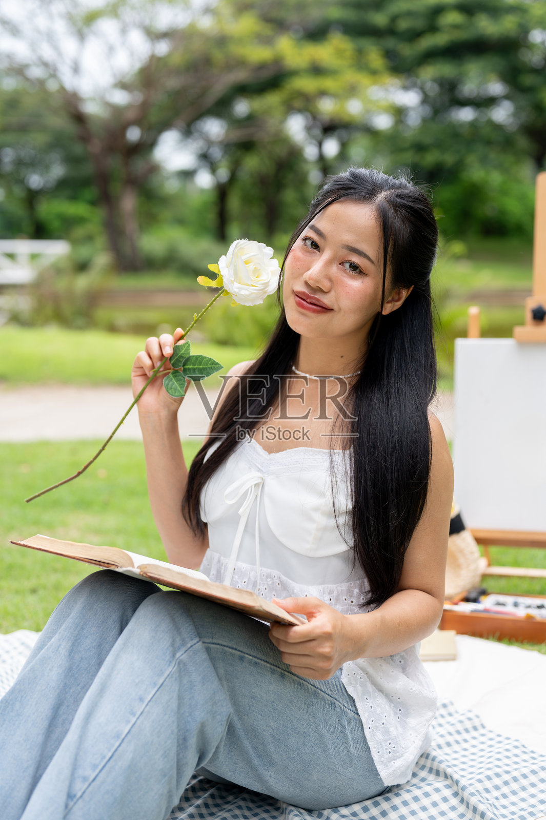 一位美丽的亚洲女性坐在公园的野餐毯上，对着镜头摆姿势微笑。照片摄影图片