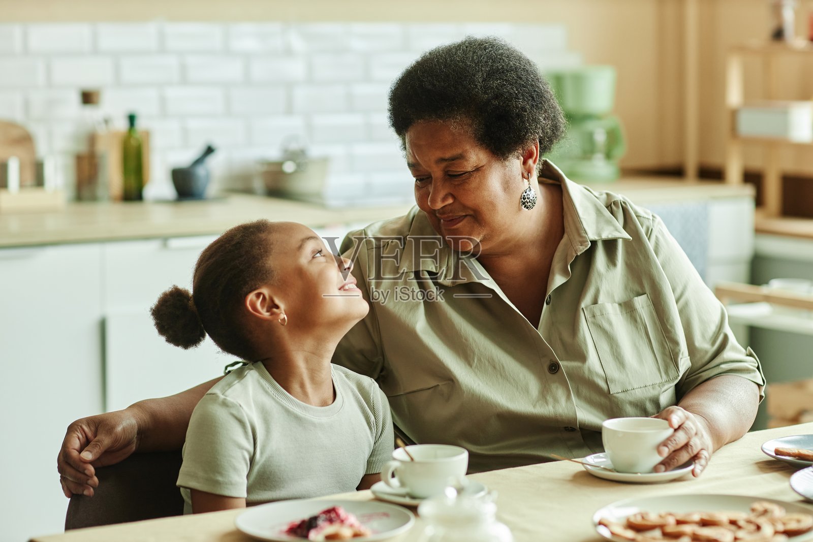 快乐的奶奶用爱的目光看着在厨房餐桌上喝茶的女孩照片摄影图片