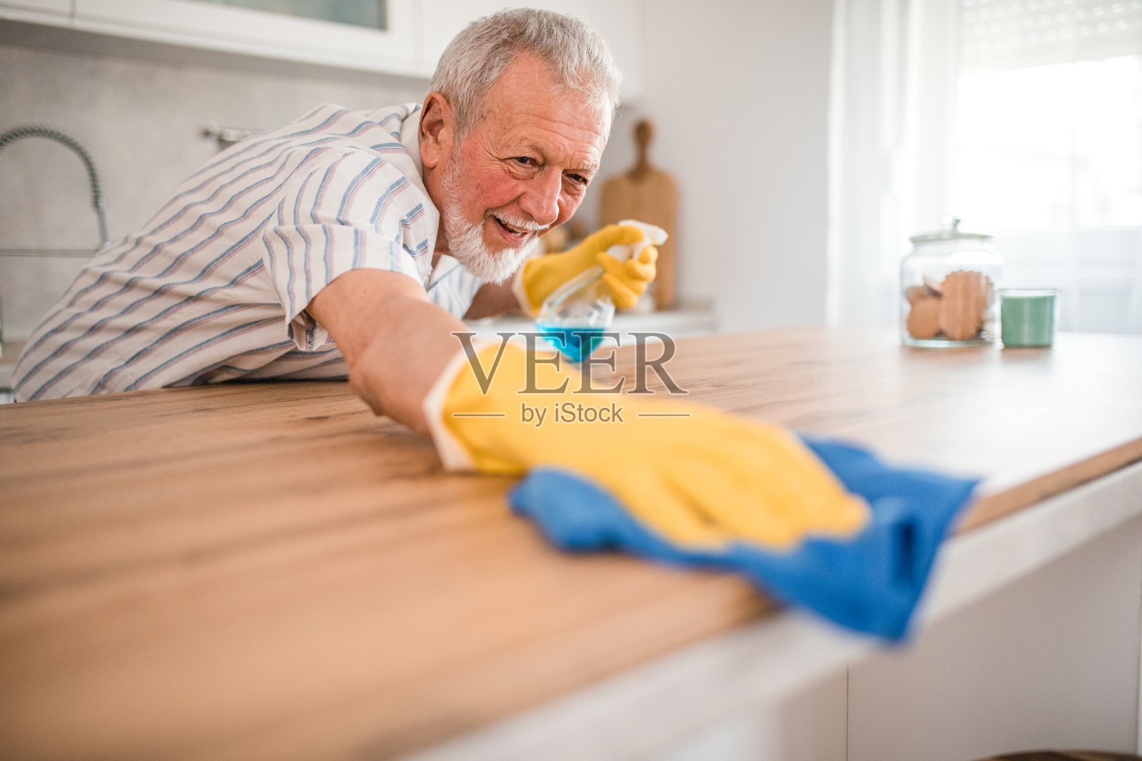 一个无忧无虑的老人带着防护手套打扫房子，擦去厨房木质表面上的灰尘照片摄影图片