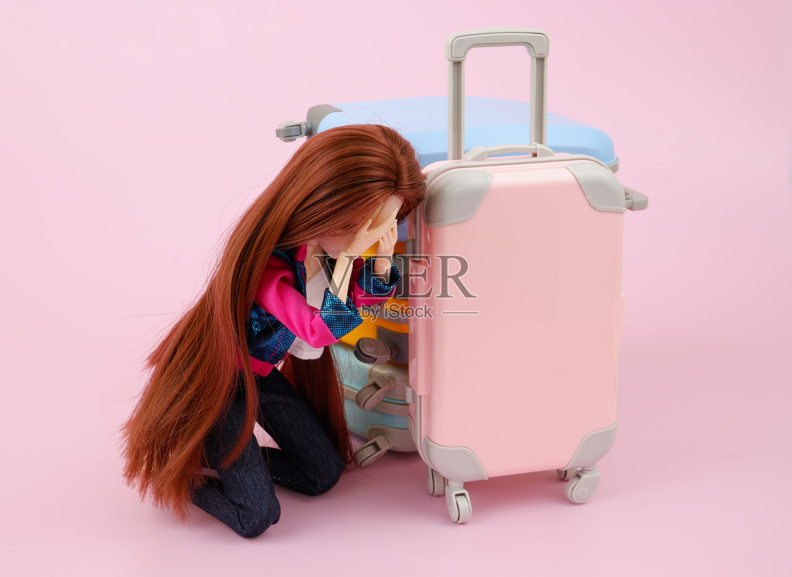 粉红色背景的普通塑料女娃娃在许多行李箱旁哭泣。照片摄影图片