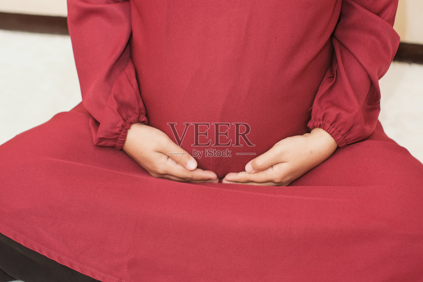 一名穿着礼服的孕妇坐在奶油色背景上，双手放在肚子上。照片摄影图片