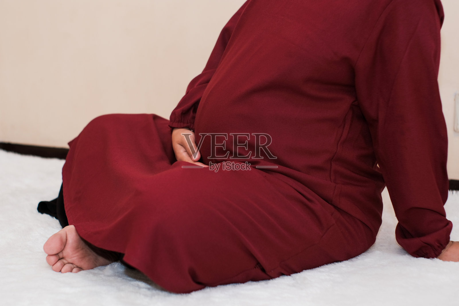 一名穿着礼服的孕妇坐在奶油色背景上，双手放在肚子上。照片摄影图片