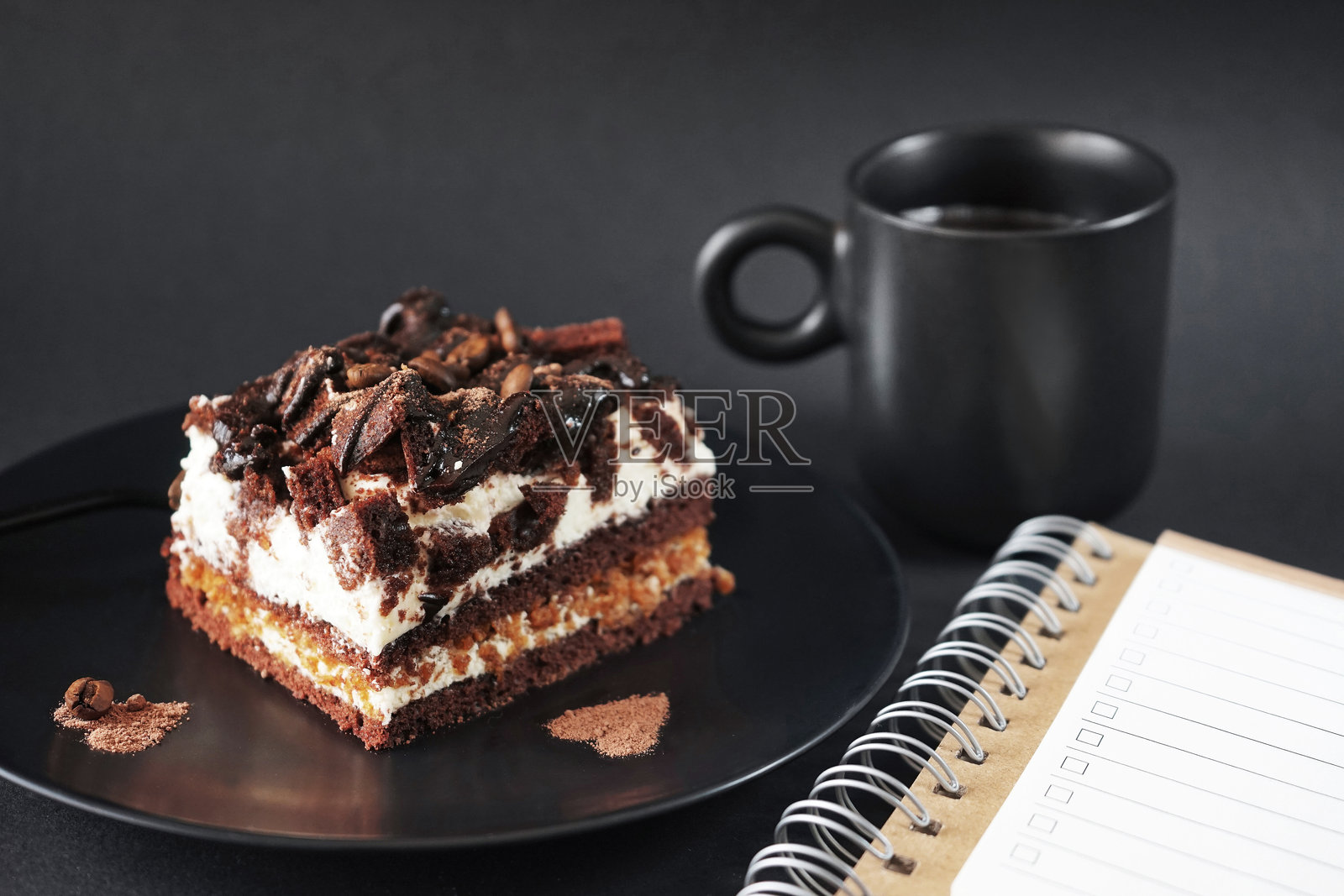 黑盘子里放着咖啡味的巧克力蛋糕，旁边是一杯咖啡和记事本照片摄影图片