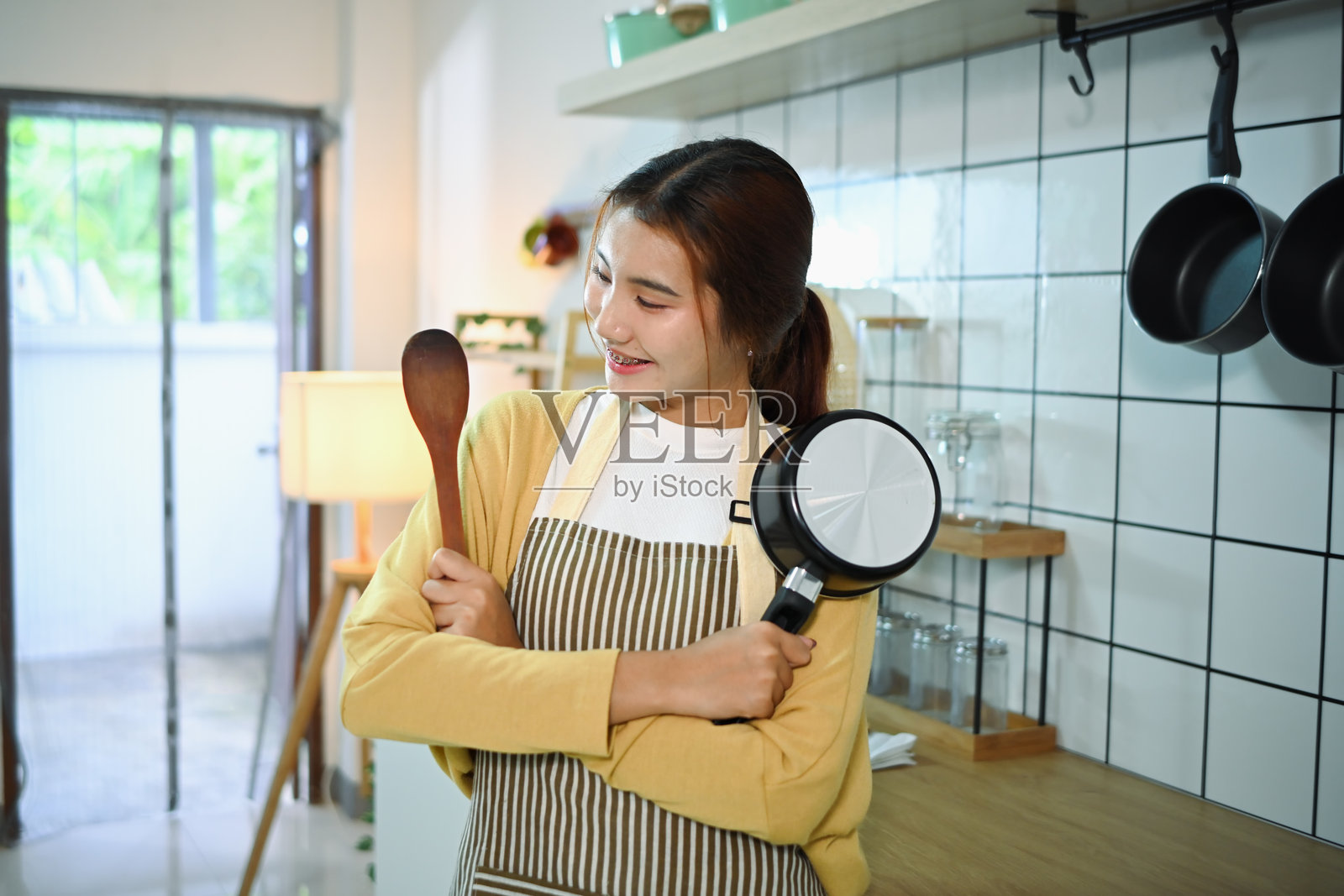 年轻女子手持木铲和锅站在厨房里的画像。居家生活和烹饪理念照片摄影图片