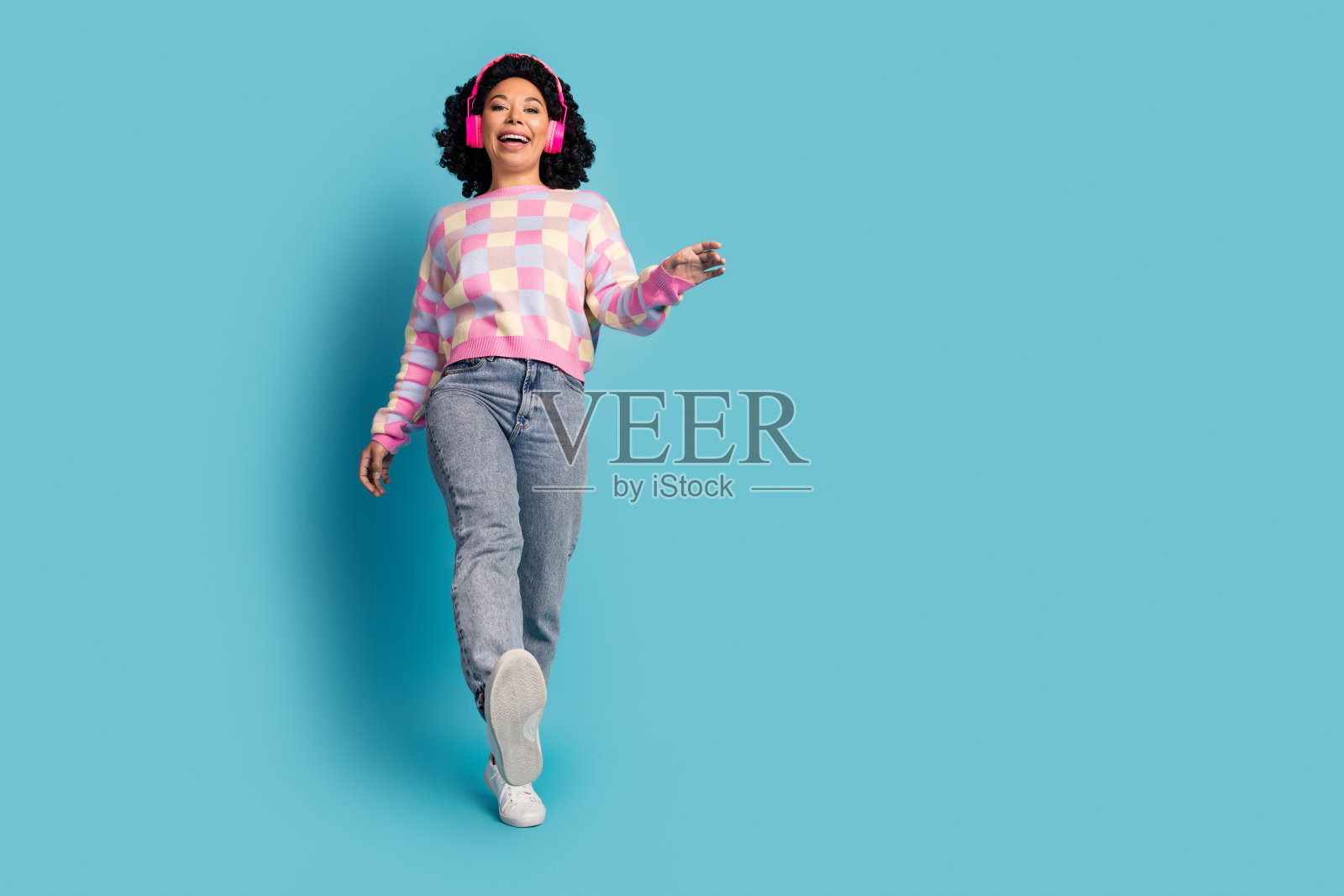 迷人的年轻女子的全身照片，戴着耳机跳舞，穿着时尚的粉红色格纹衣服，孤立在蓝色背景上照片摄影图片