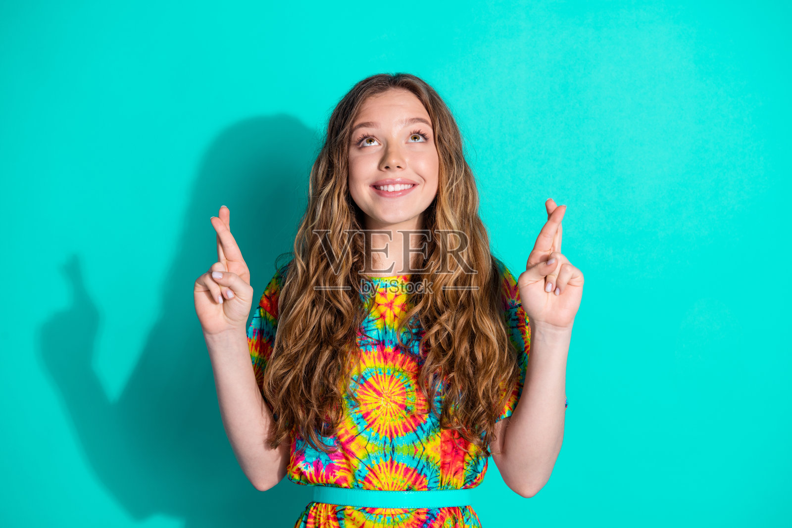 漂亮的十几岁女孩的照片，交叉手指许愿，仰望空旷的空间，穿着时髦的彩色服装，孤立在海蓝宝石的背景上照片摄影图片