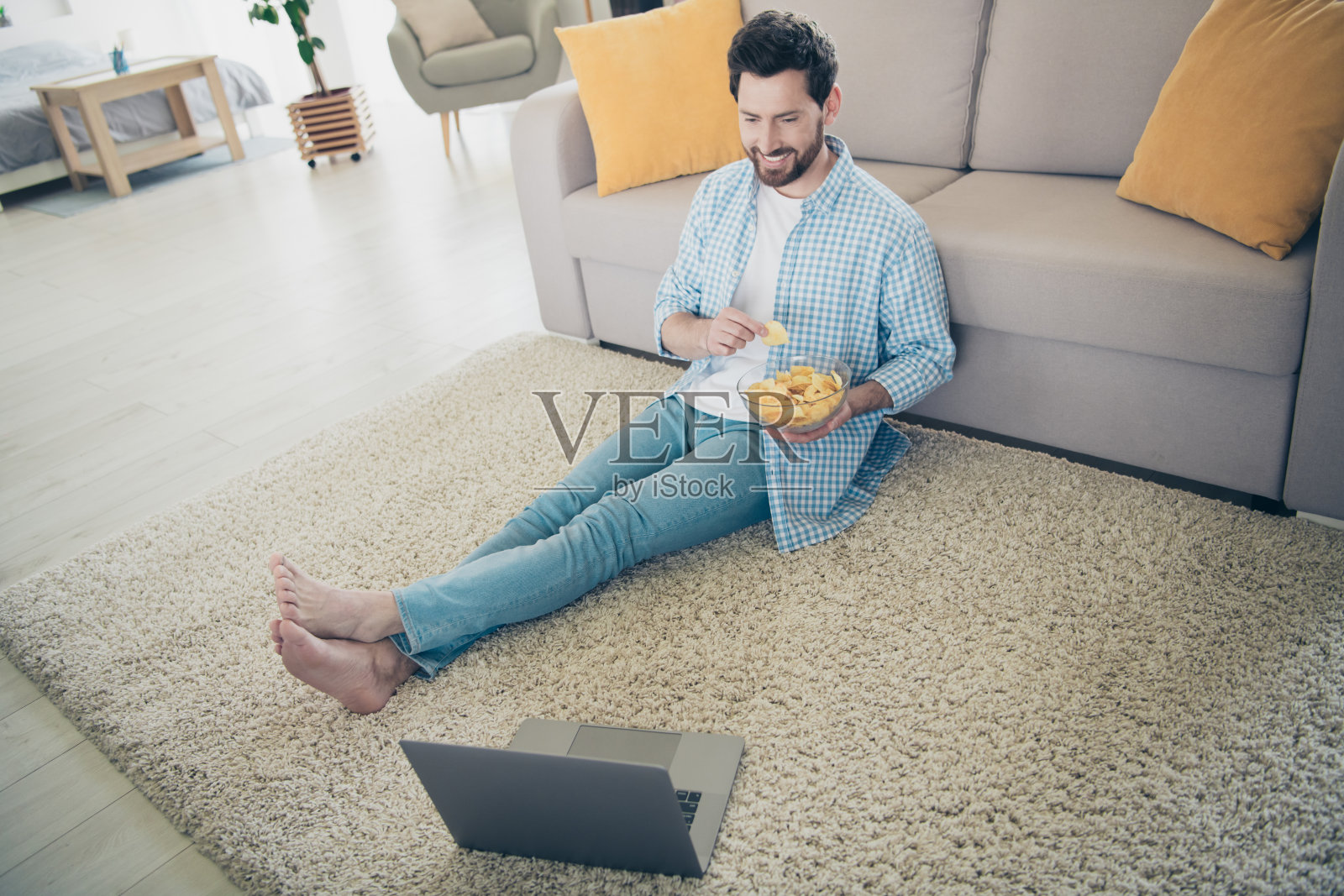 大胡子的成年男子独自呆在家里的新房子里，在客厅里，坐在地板上吃薯片，看喜剧照片摄影图片