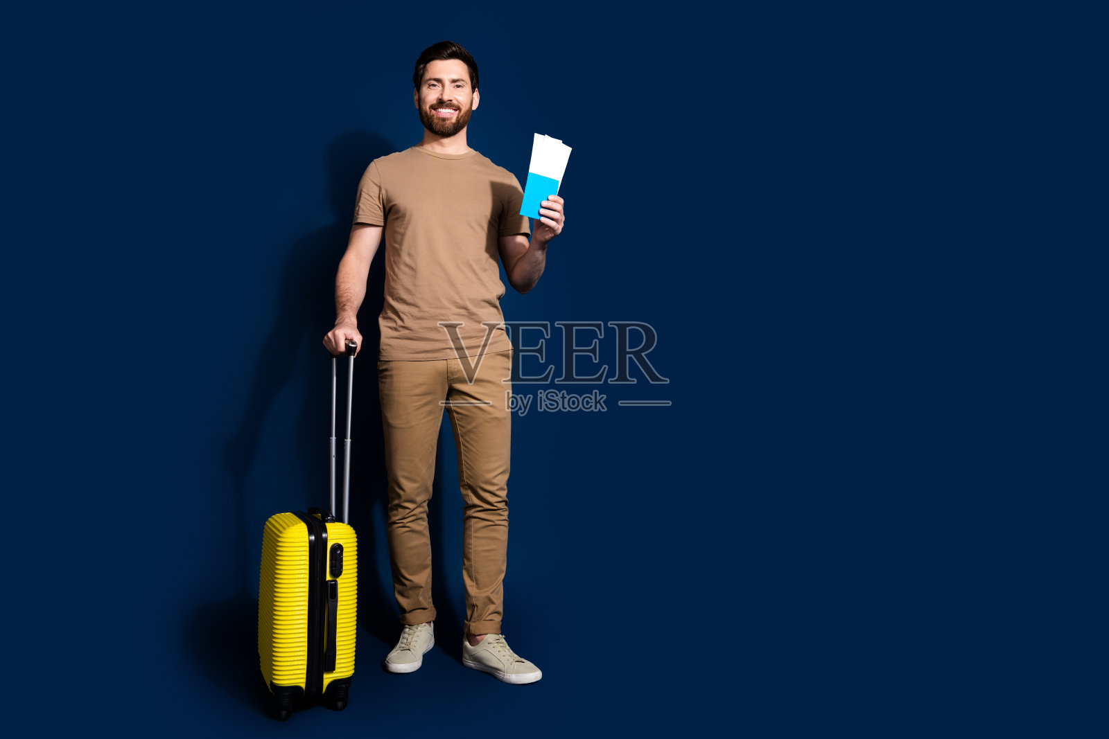 年轻帅哥的照片肖像，深色头发的模特，穿着米色t恤，拿着行李和机票去机场，在深蓝色的背景上孤立起来照片摄影图片