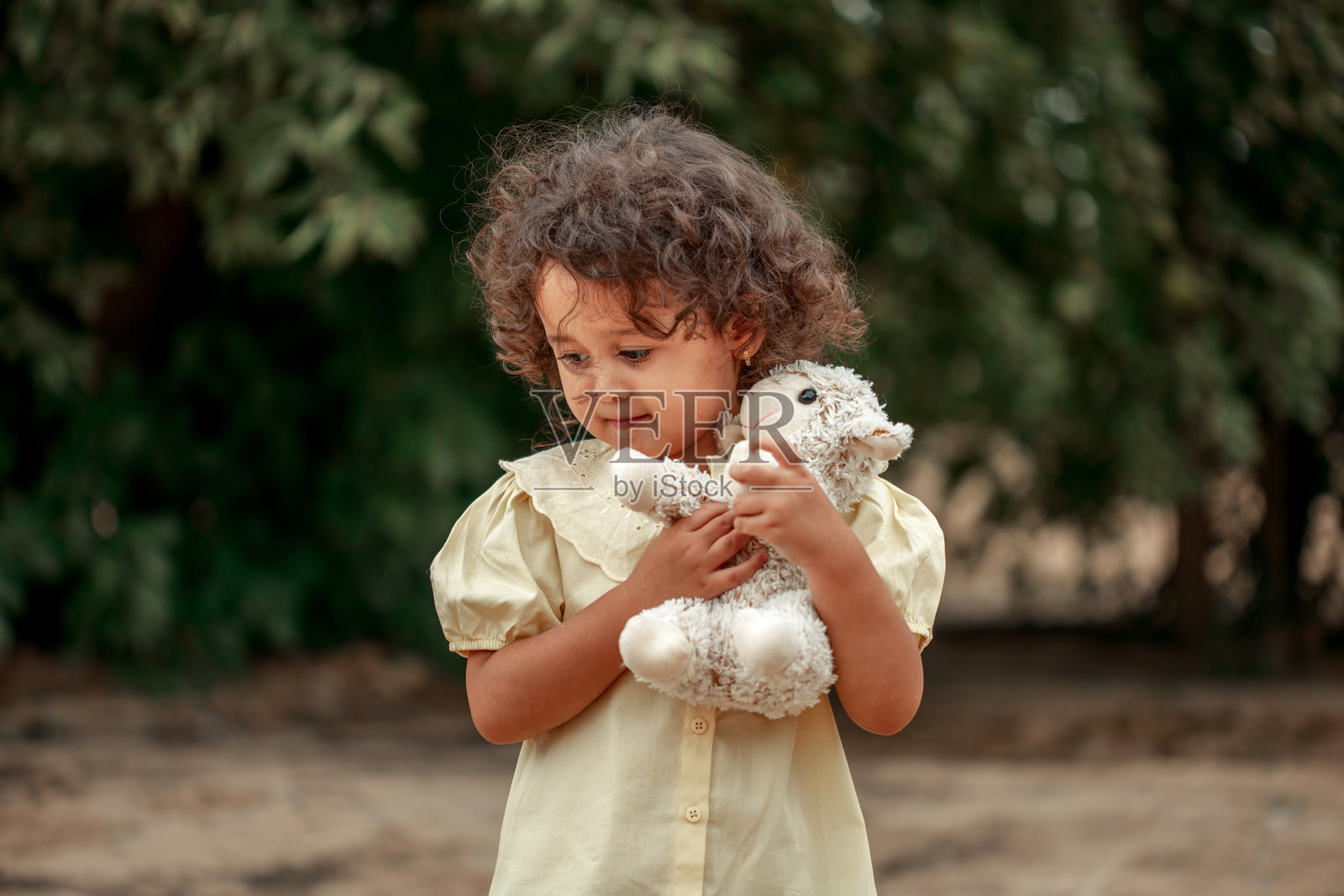 肖像。一个穿着米色连衣裙的小卷发女孩站在那里，手里拿着一个玩具。照片摄影图片