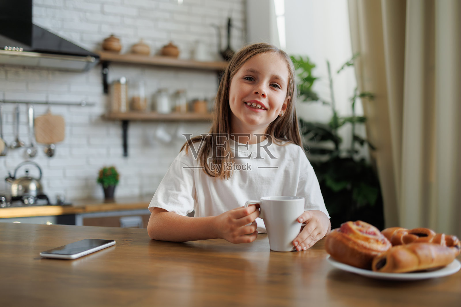 微笑的青春期前女孩在厨房里拿着杯子，旁边是馅饼和智能手机照片摄影图片