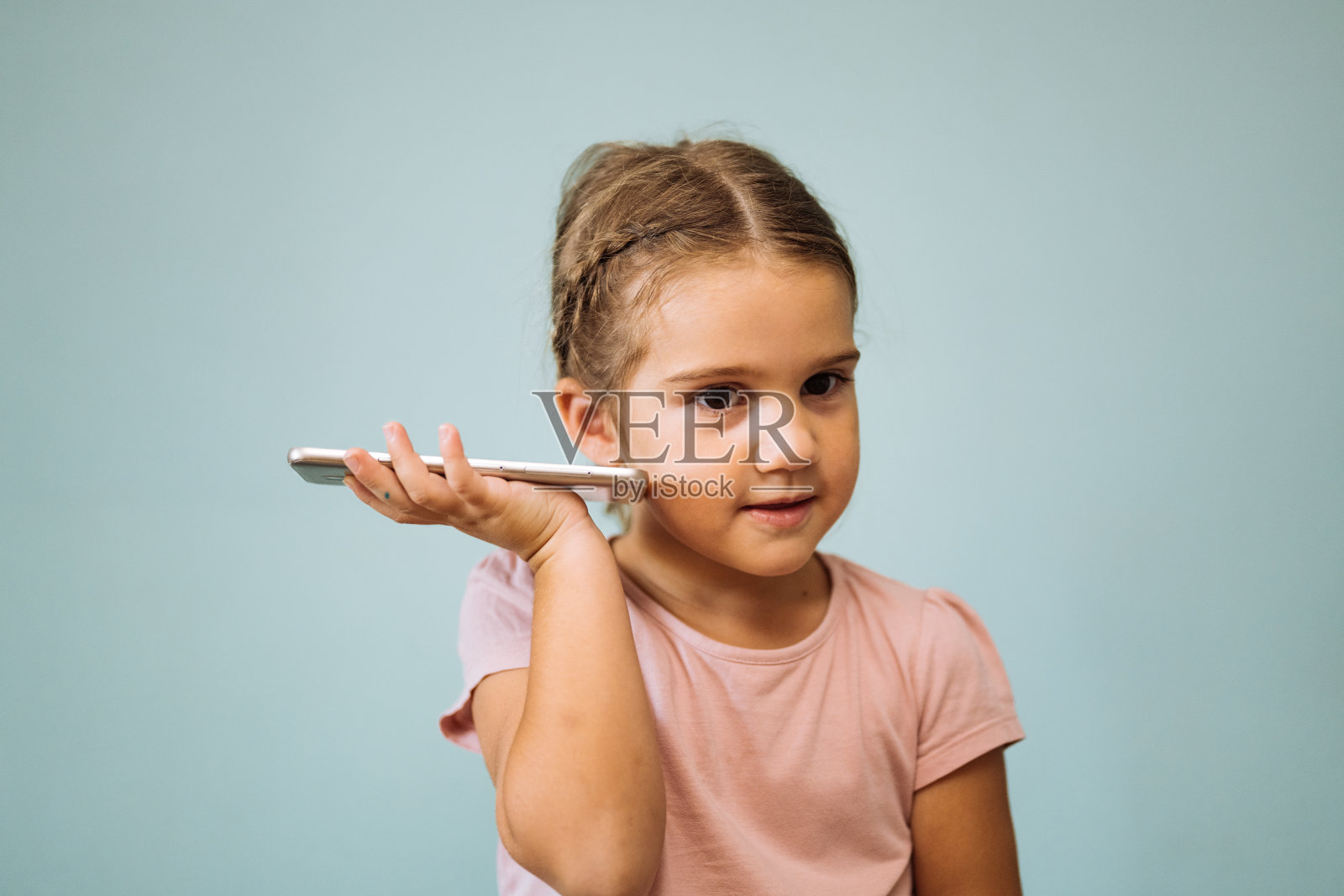 一个年轻的女孩，穿着中性浅蓝色背景的粉色衬衫，把智能手机放在耳边，好像在说话。照片摄影图片