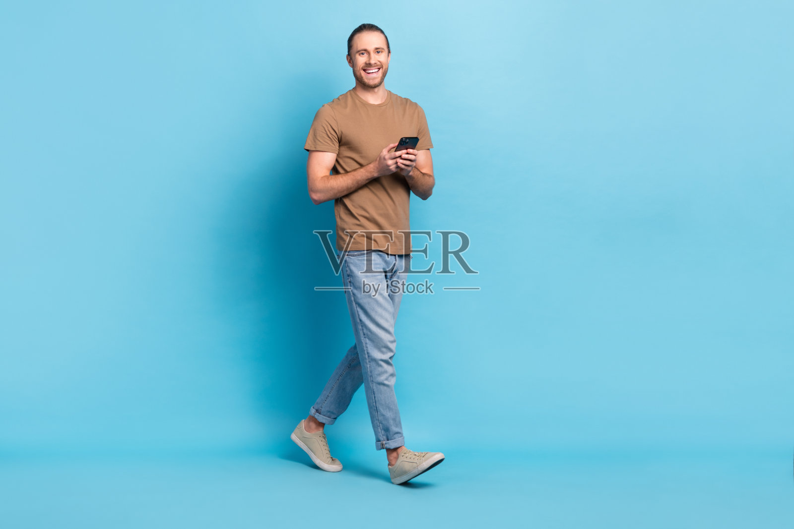 全长照片正面漂亮的家伙穿着米色t恤打字现代装置行走空旷的空间孤立的蓝色背景照片摄影图片