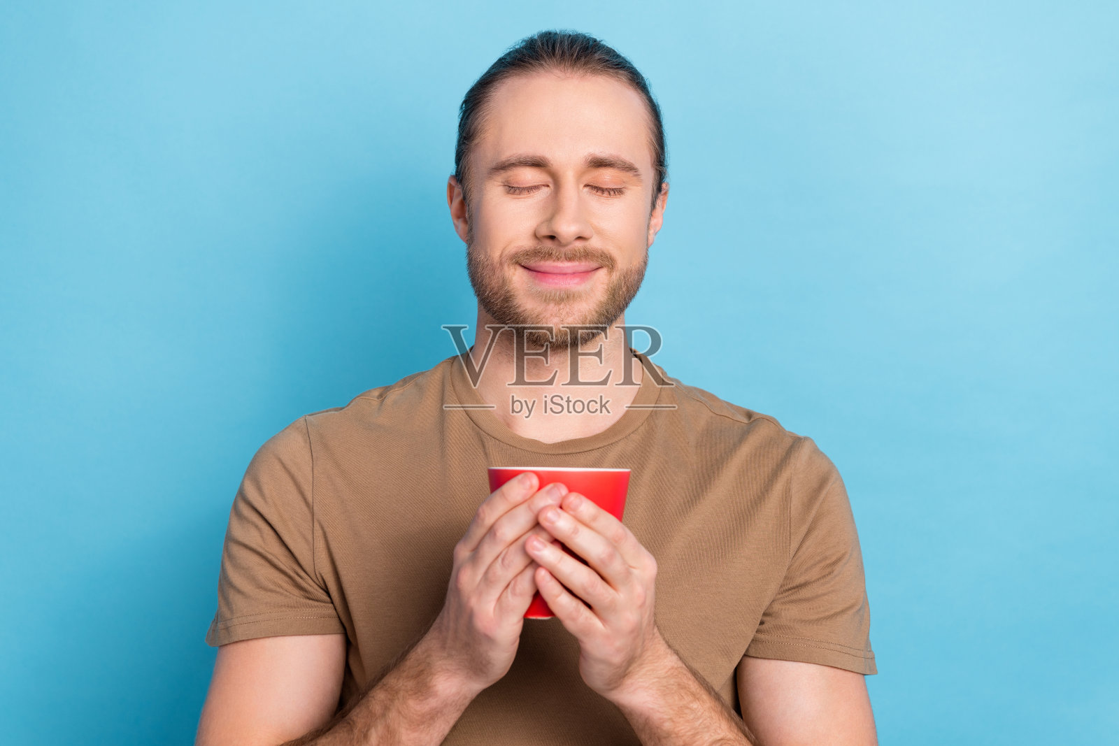 照片开朗轻松英俊的男人尾巴发型穿着米色t恤拿着杯子享受早晨的时间孤立的蓝色背景照片摄影图片