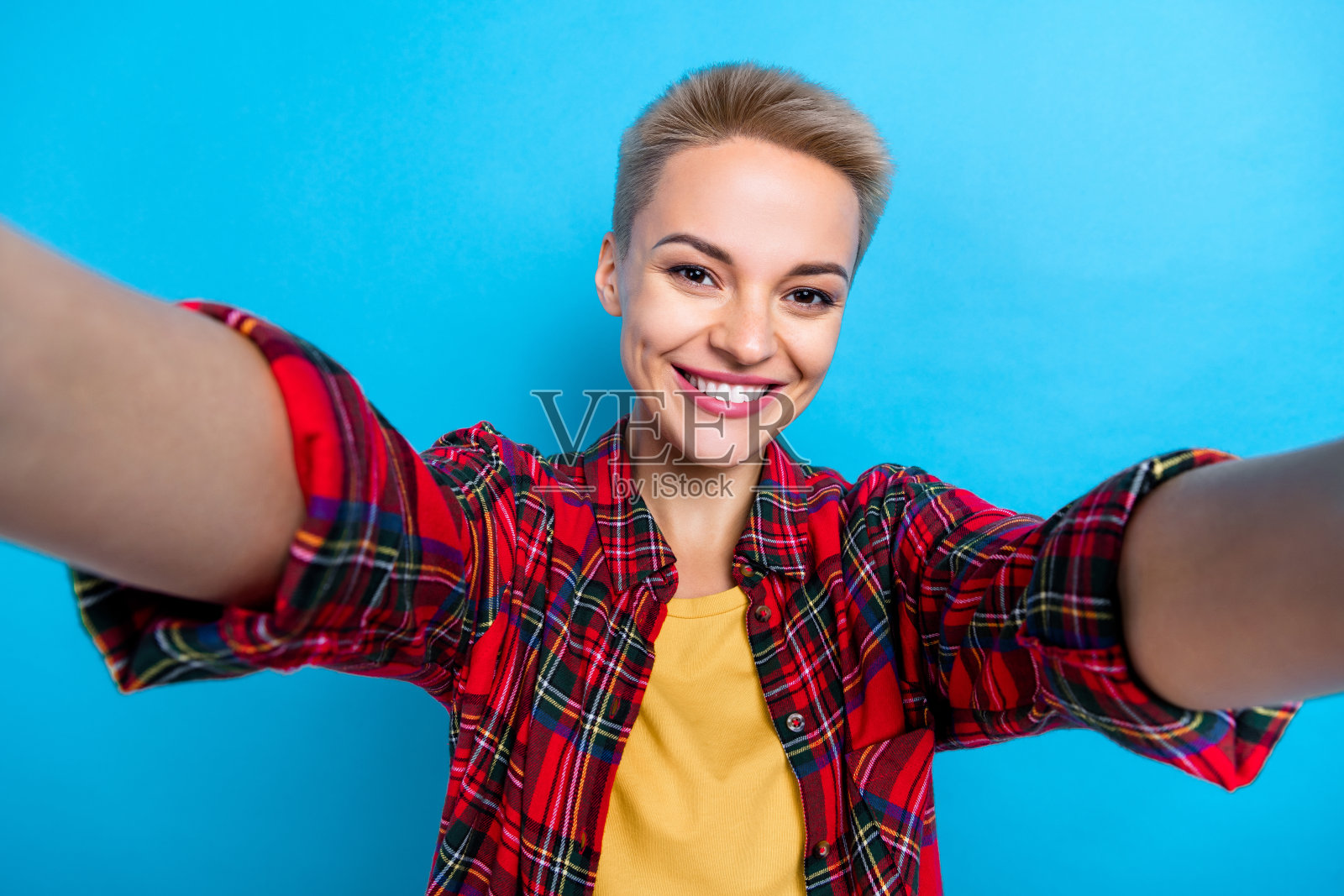 漂亮可爱的短发女人的肖像，穿着格子衬衫，拿着相机在蓝色背景下自拍照片摄影图片