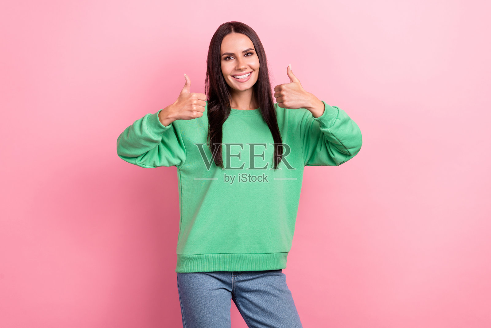 一个乐观满意的女人，留着长发，穿着绿色运动衫，在粉红色的背景上竖起大拇指照片摄影图片