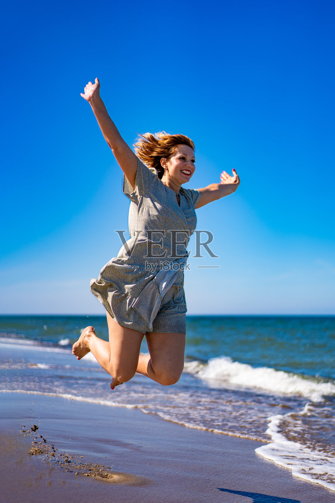 夏日的海边，美丽的沙滩上，年轻漂亮的女子穿着连衣裙，高举双臂，欢快地跳着。生命的力量和乐趣照片摄影图片