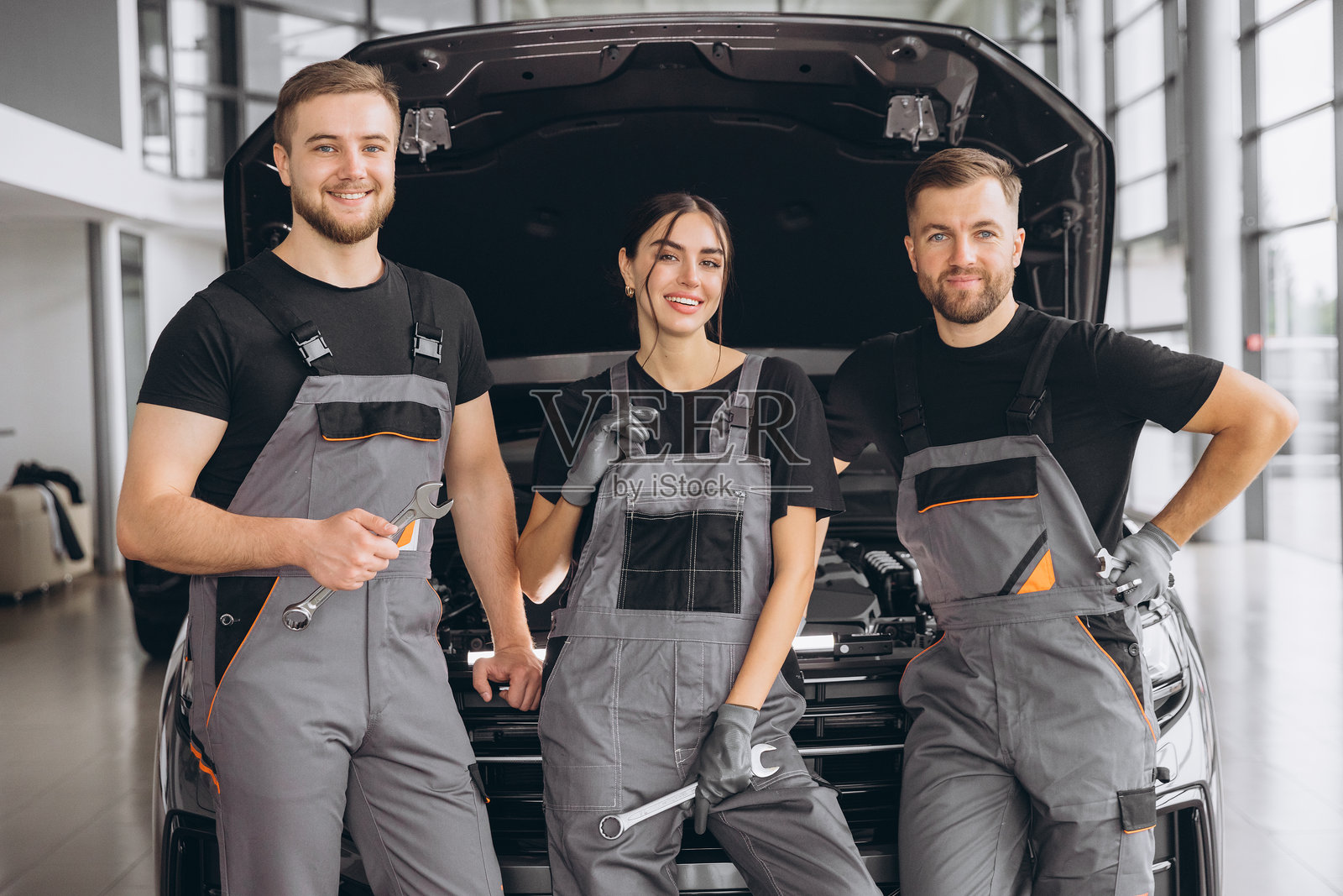 三个面带微笑的工人，机械师两男一女穿着灰色制服站在打开的引擎盖，车库汽车商店，汽车机械师维修客户的汽车汽车在维修服务。照片摄影图片