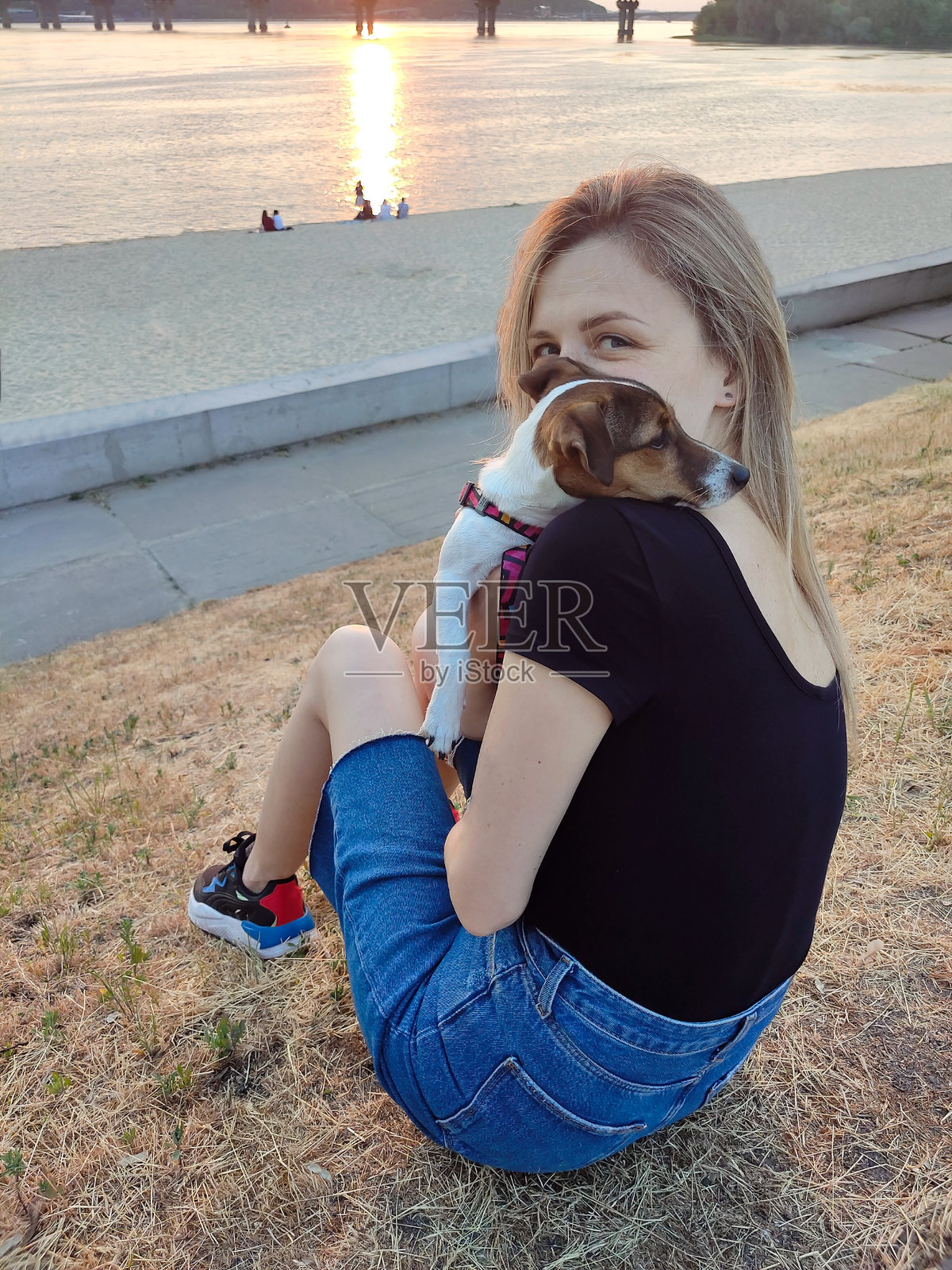 夕阳西下，一个金发女孩抱着一只杰克罗素梗狗坐在河岸上照片摄影图片