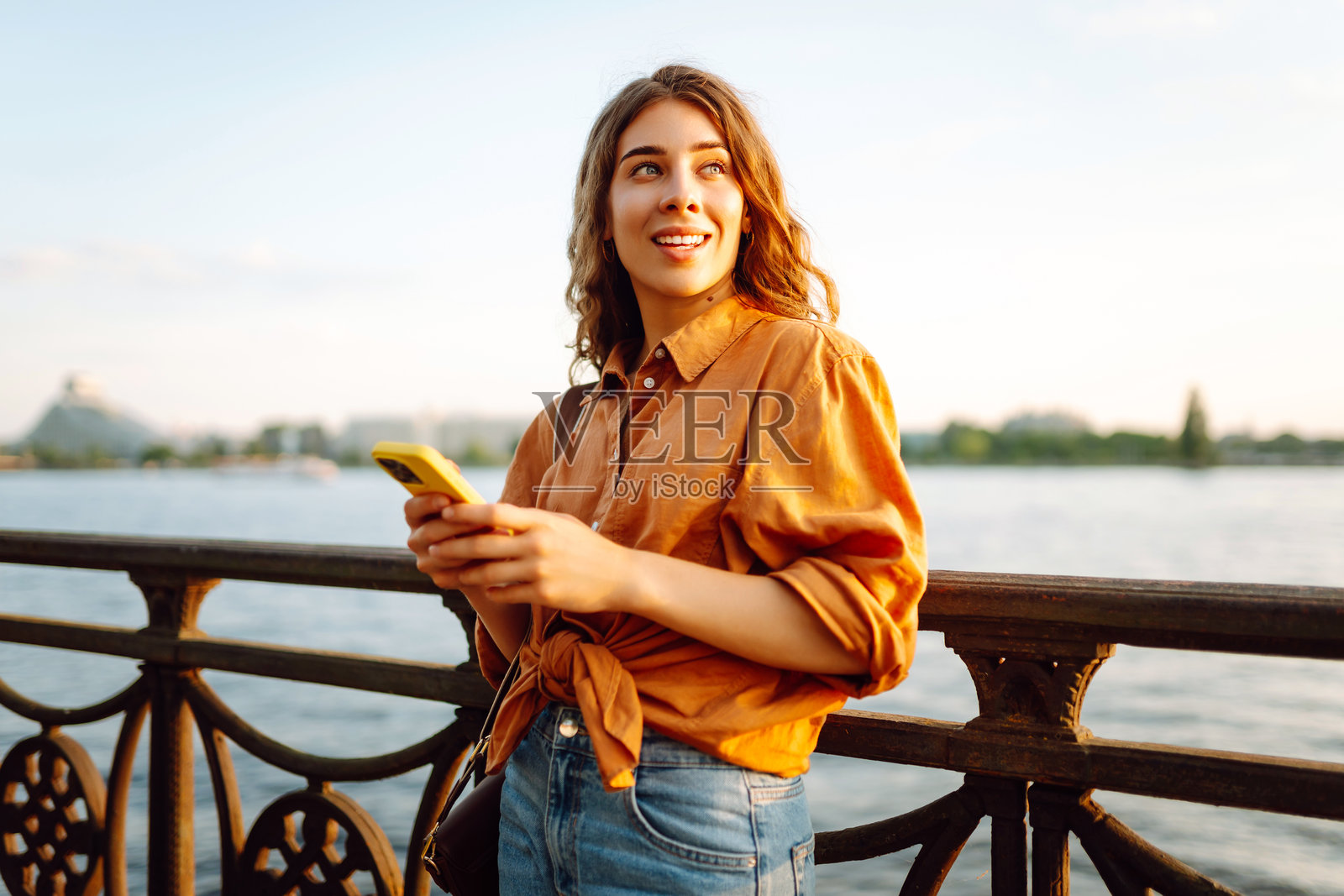 日落时分，一名年轻女子在桥上用智能手机微笑。照片摄影图片