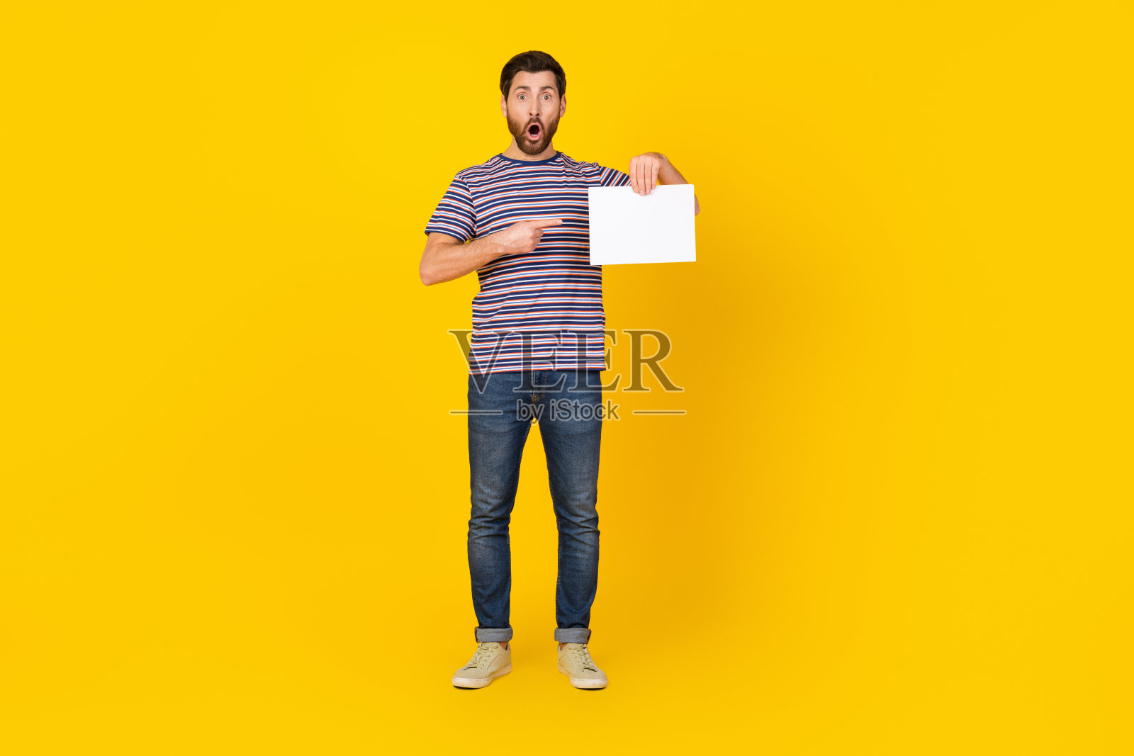 一名身穿条纹t恤的男子震惊地用手指向黄色背景上的广告牌空白区域照片摄影图片